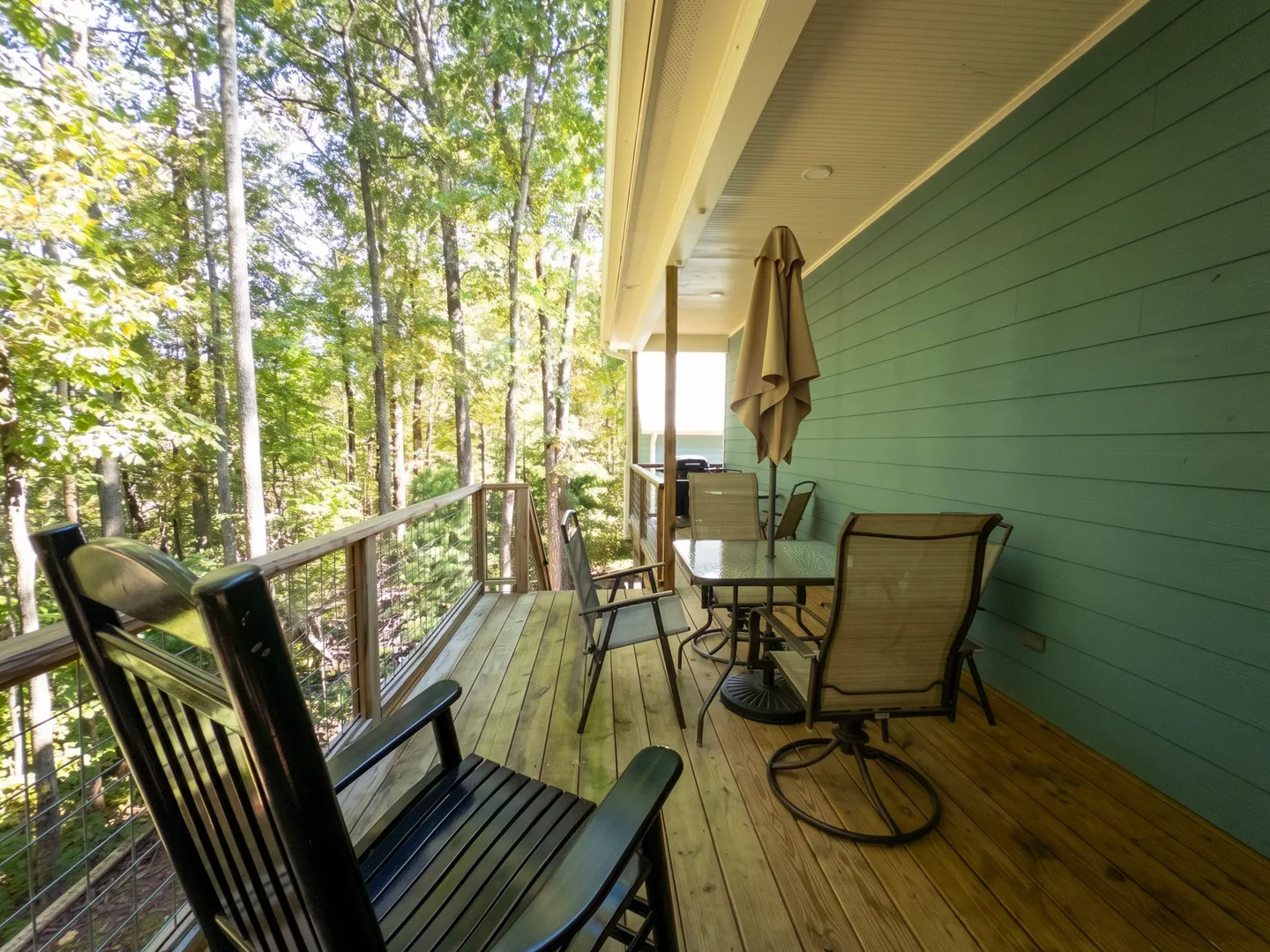 A covered wooden deck furnished with a black rocking chair, dining table, chairs, and patio umbrella, overlooking surrounding trees.