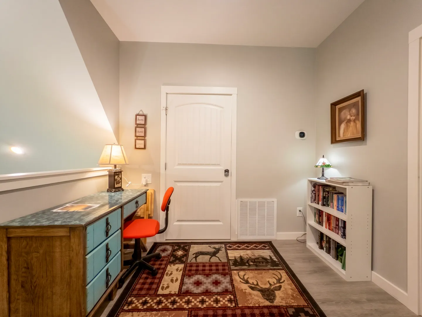 Workspace area featuring a desk with a lamp and orange chair, a white door, a bookshelf, and a rustic-themed area rug.