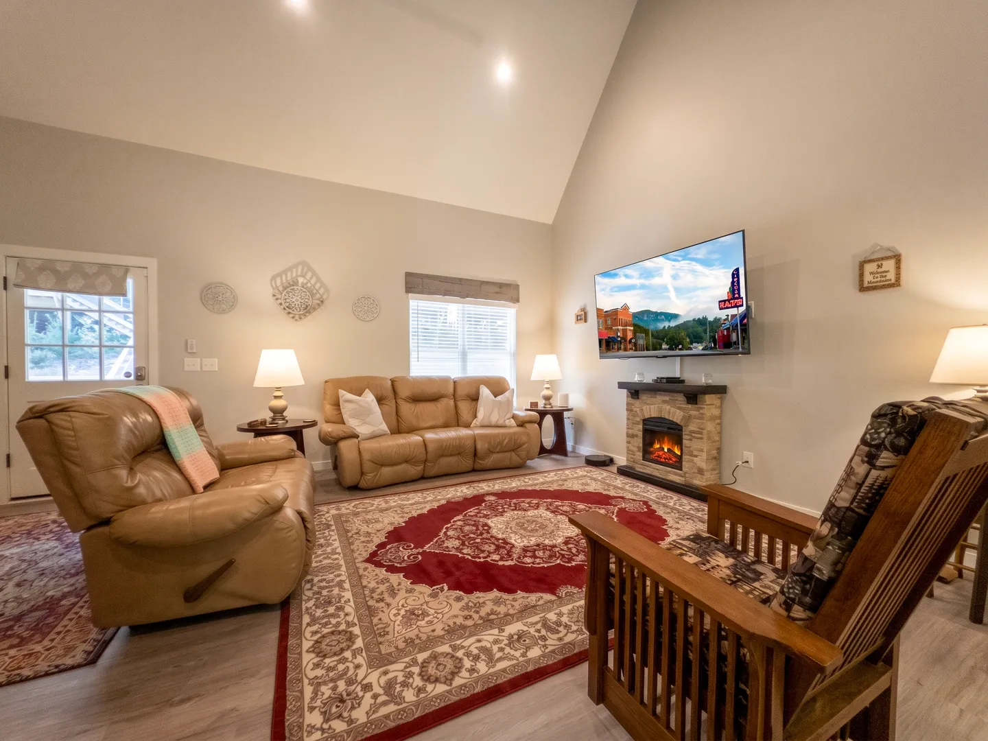 Living room featuring vaulted ceilings, a leather sofa, recliner, patterned arm chair, area rug, and an electric fireplace with a wall-mounted TV above.
