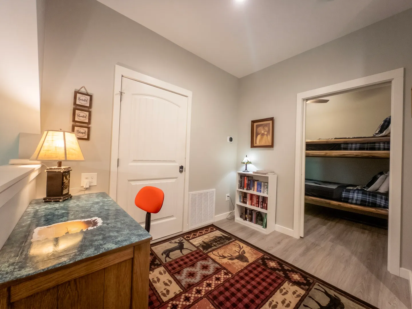 An office or study nook with a wooden desk, orange chair, rustic rug, and a view into a bunk bedroom.