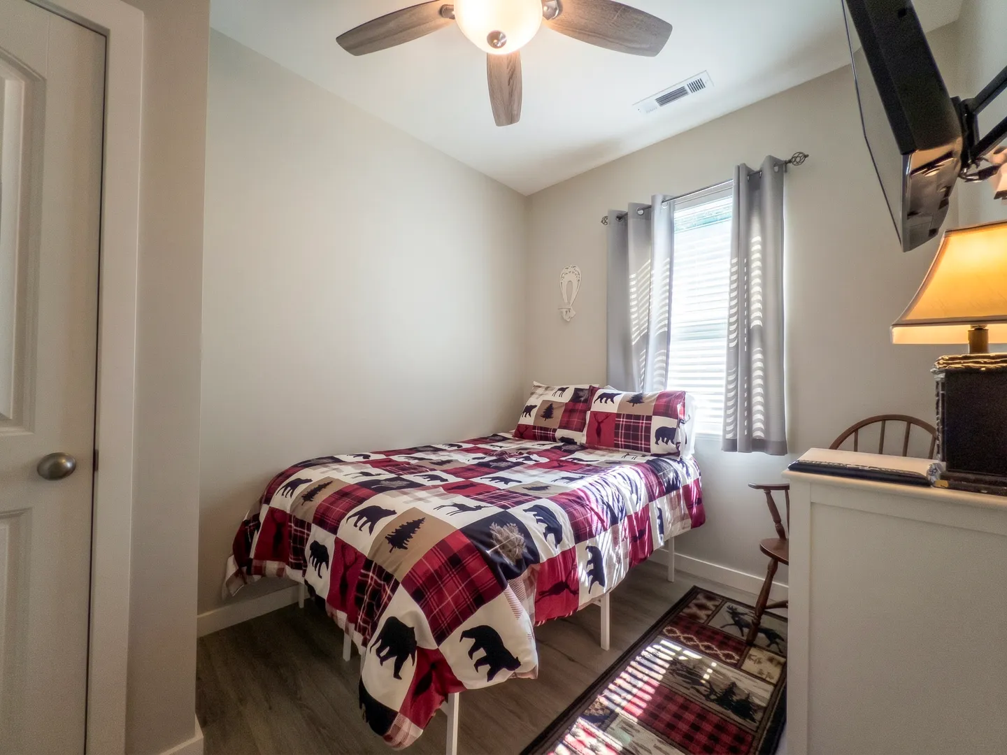 Bedroom with a bed made with a rustic bear-and-plaid quilt, a ceiling fan, a window with grey curtains, and a television.