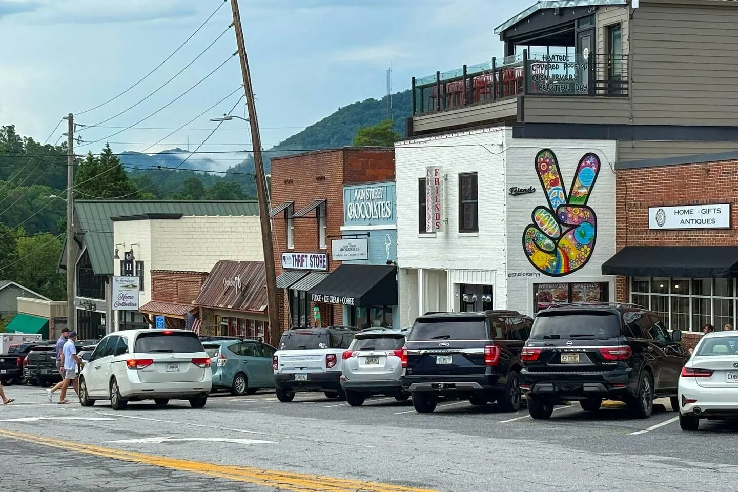 Street view of local downtown shops and parked cars with forested mountains in the background under an overcast sky.