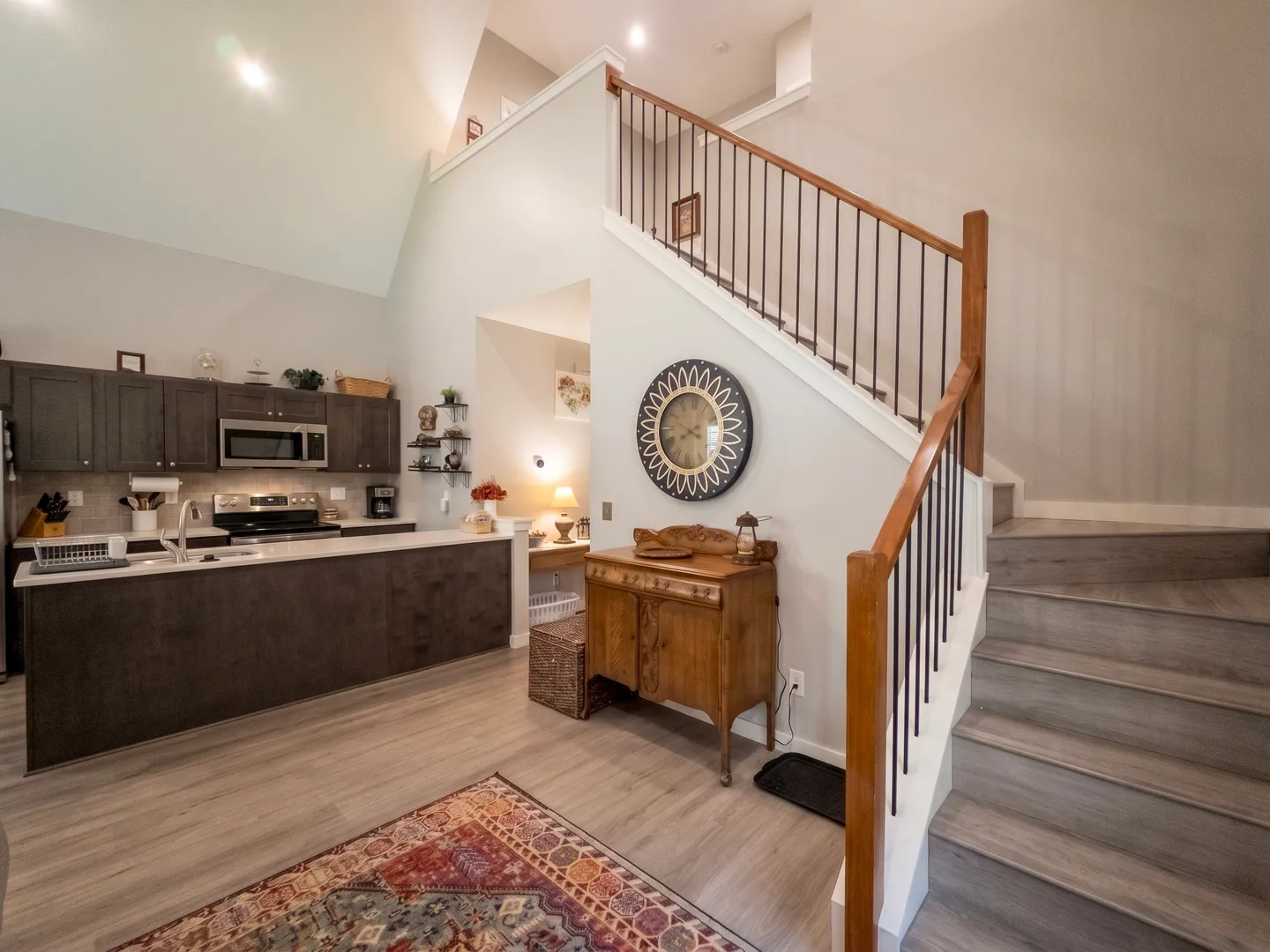 Open-concept living space showing wood-look stairs with black metal spindles next to a kitchen with dark cabinetry and vaulted ceilings.