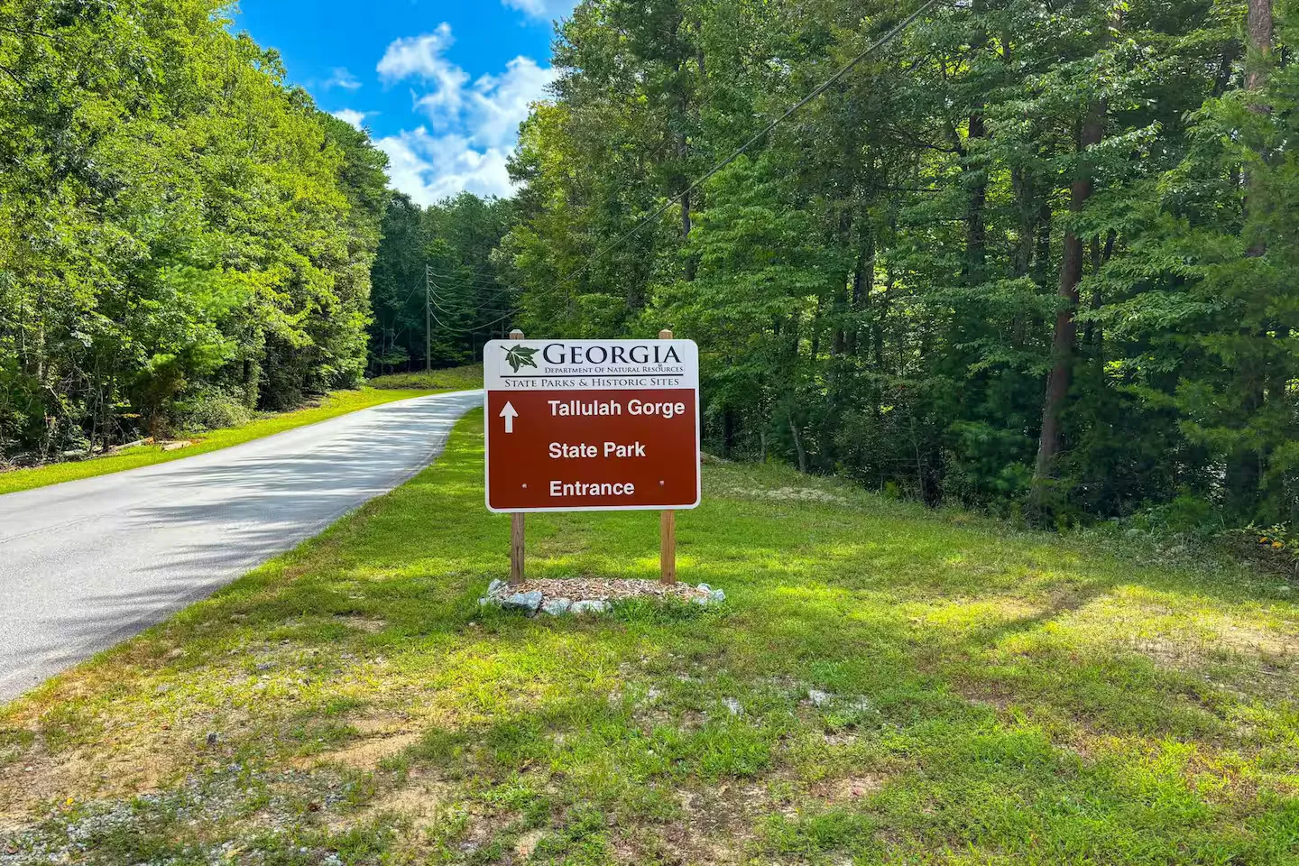 Brown Georgia State Parks sign pointing straight ahead to Tallulah Gorge State Park Entrance alongside a paved road lined with dense trees.