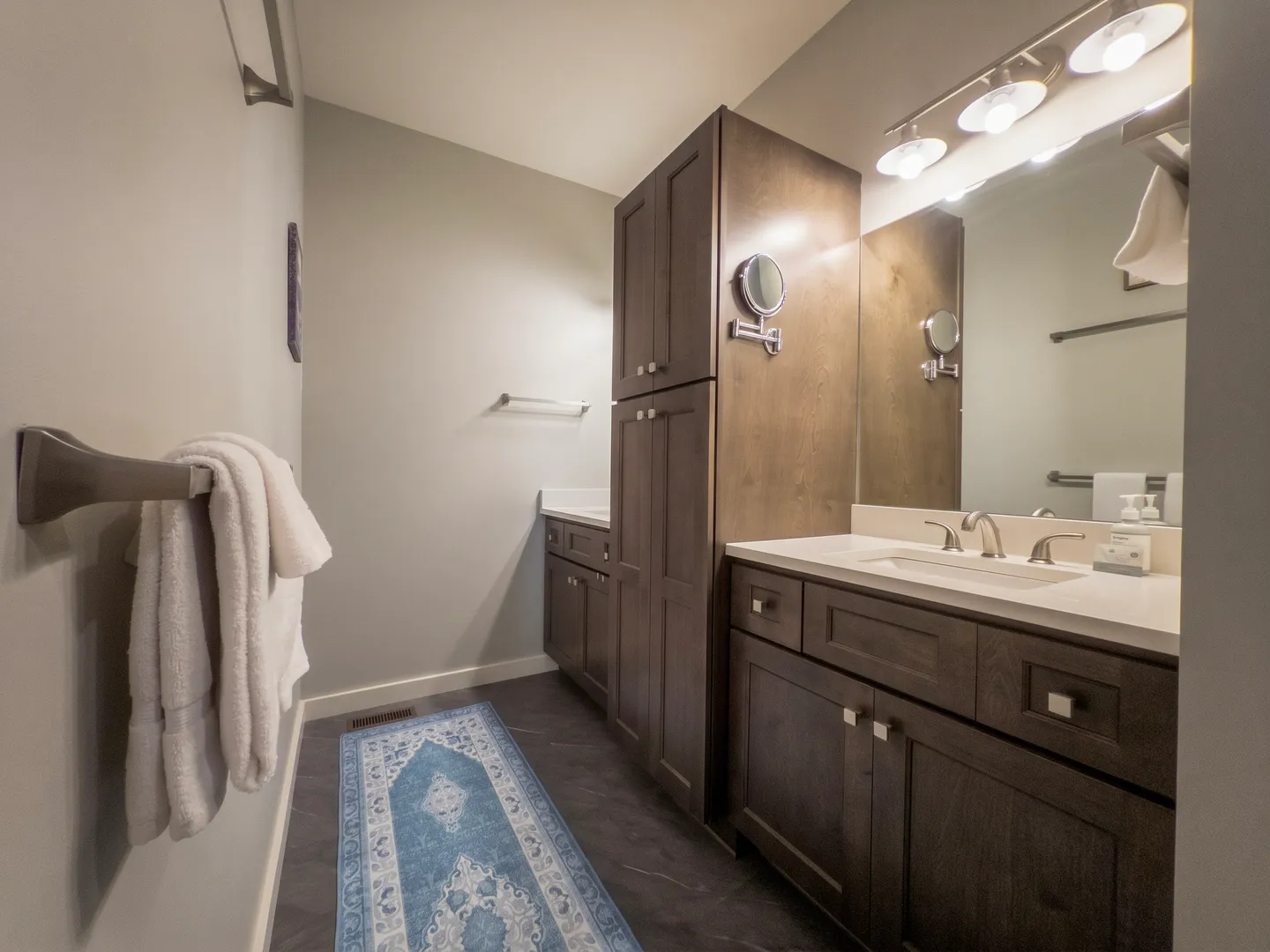 Bathroom featuring dark wood vanities and tall storage cabinet with light countertops, illuminated mirror, and a blue patterned runner rug.