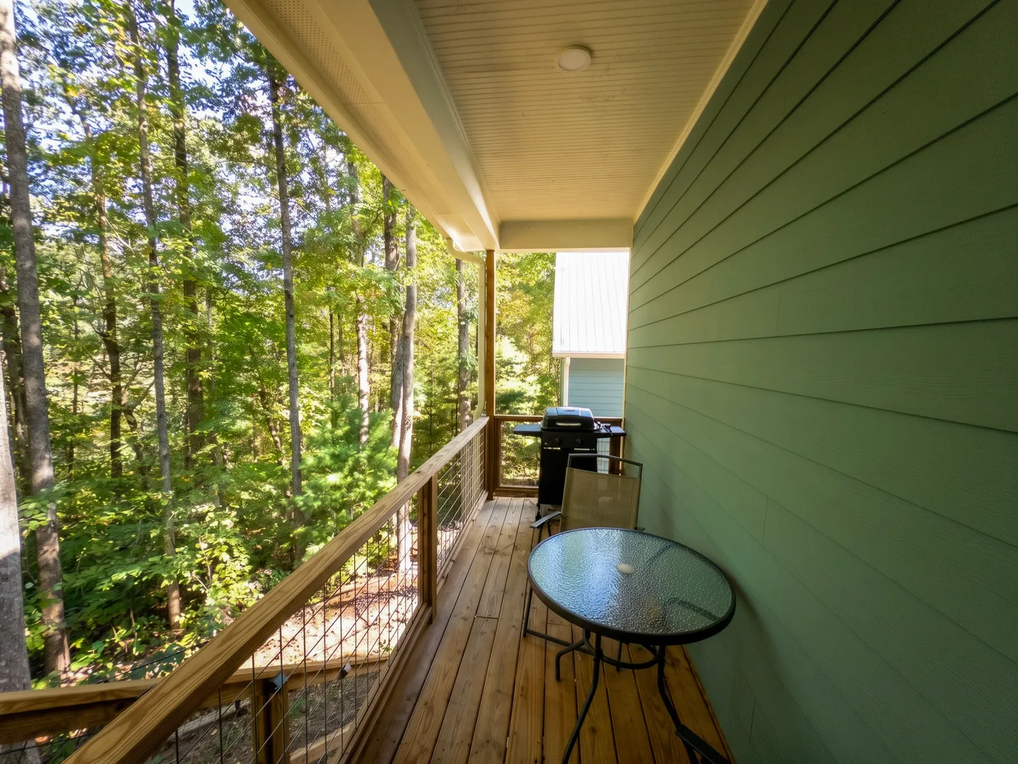 A covered wooden deck with a small round glass-top table, patio chair, and a grill, overlooking lush green woods.