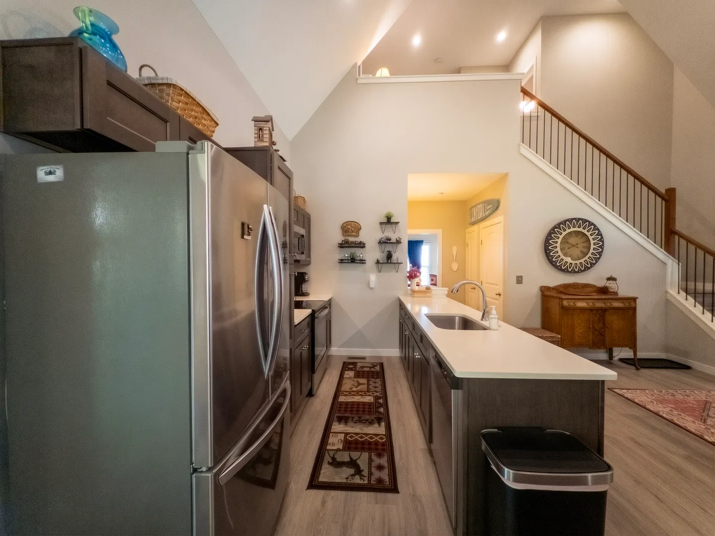 Kitchen with dark cabinets, stainless steel refrigerator and dishwasher, long island with sink, and high vaulted ceiling next to a staircase.