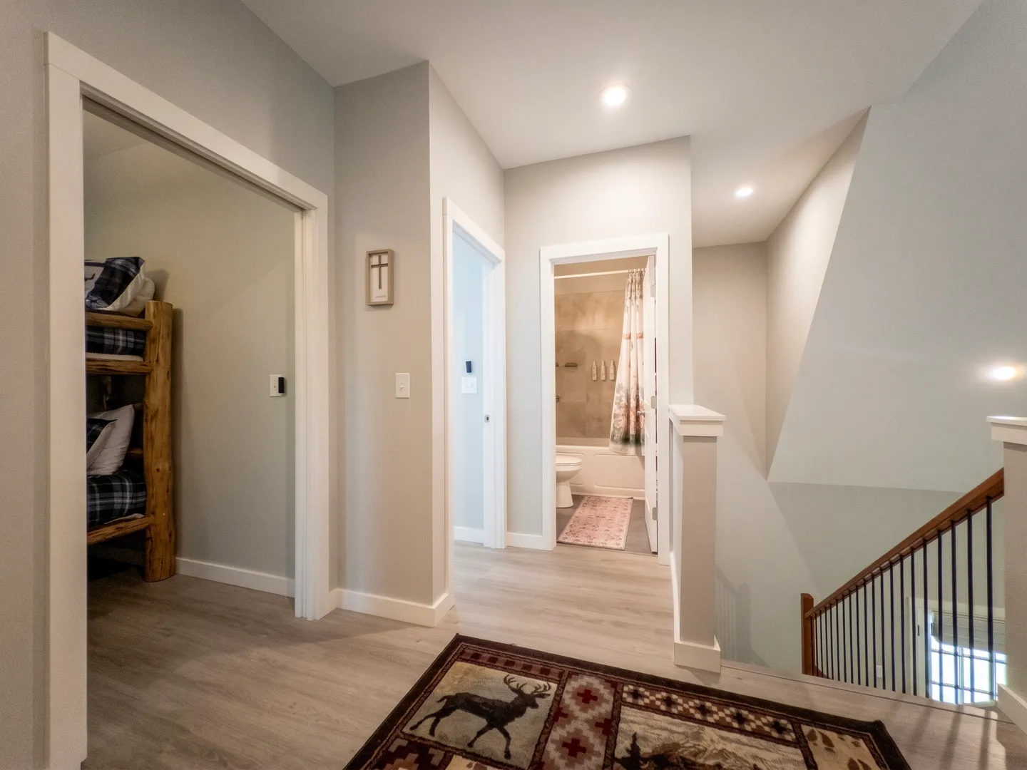 Upstairs hallway with light wood-style flooring, a staircase railing, and open doors to a bunk bedroom and a bathroom.