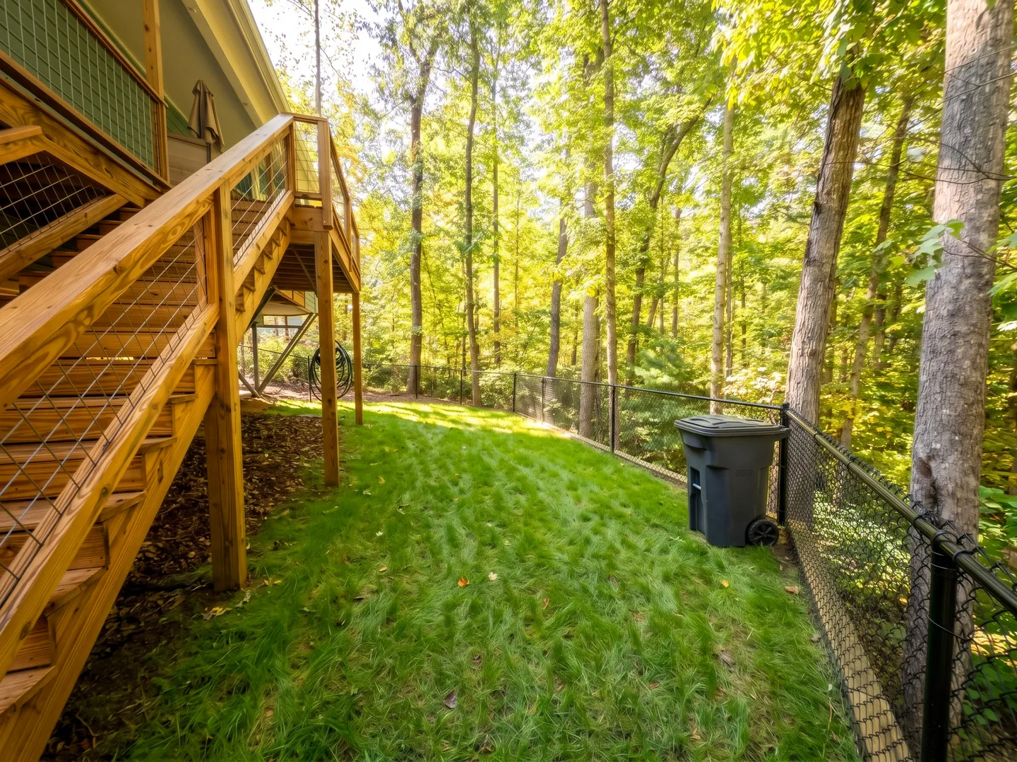 A fenced, grassy yard beneath a wooden deck staircase leading toward dense green woods.