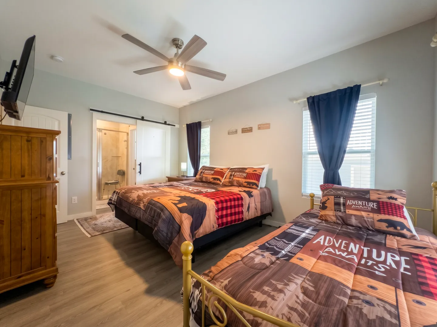 Bedroom featuring two beds with rustic bear-patterned comforters, light wood-look flooring, a ceiling fan, and an ensuite bathroom visible through a sliding barn door.