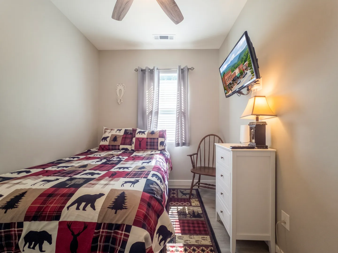 Bedroom with a single bed featuring a rustic bear and plaid quilt, a white dresser, a wall-mounted TV, and a window.