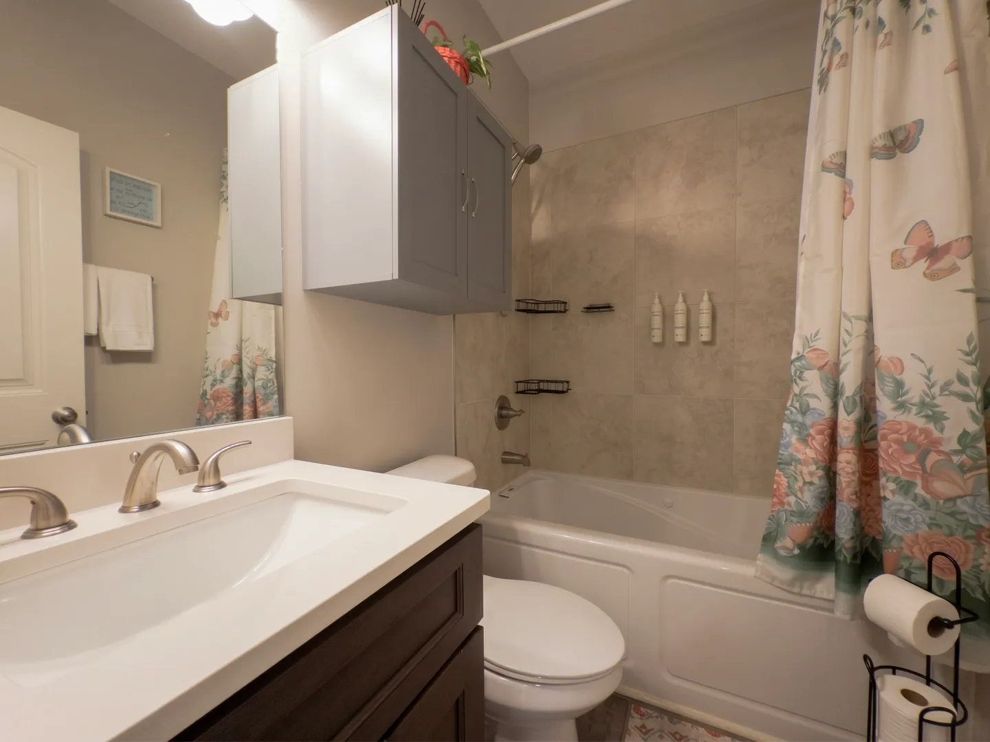 Bathroom showing a white vanity sink in the foreground, toilet, and tiled bathtub-shower combination with a floral curtain.