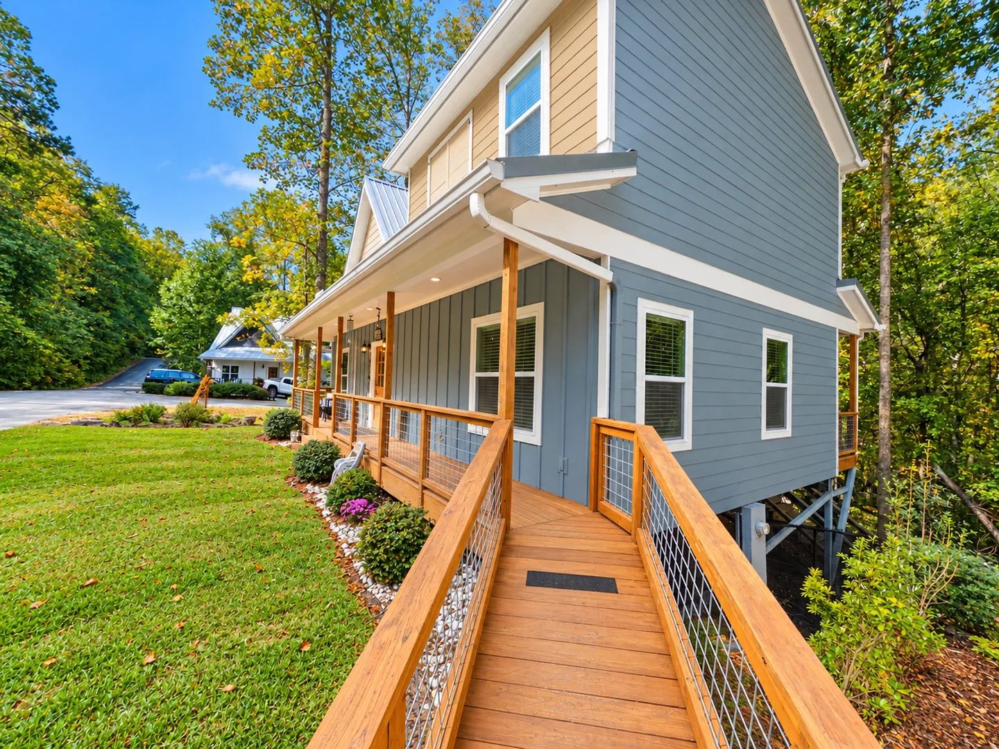 A wooden ramp with wire-mesh railings leads to the covered front porch of a blue, two-story house surrounded by a grassy lawn and trees.