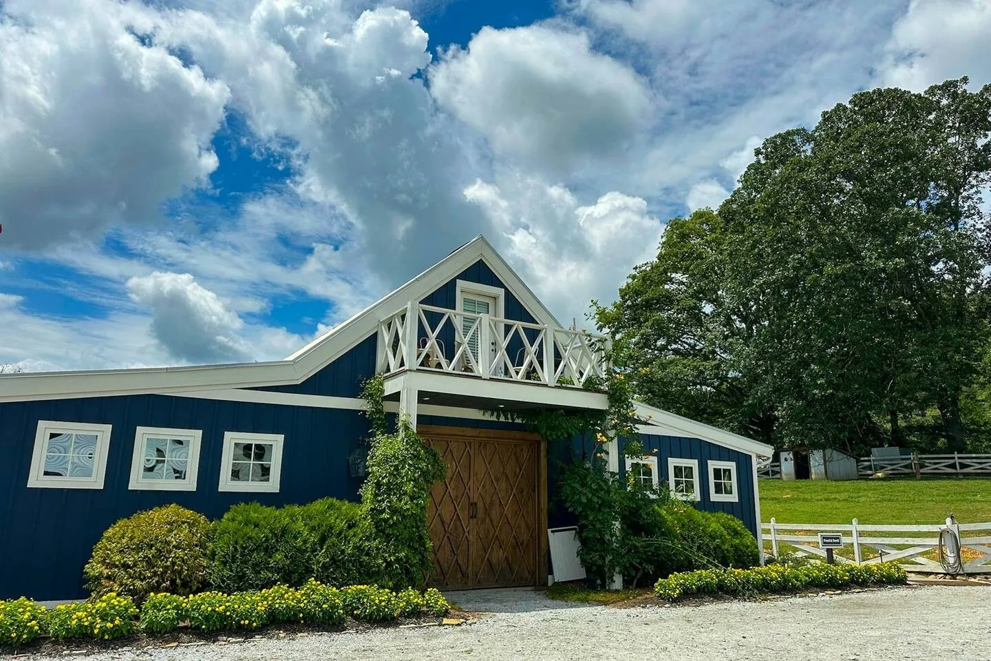 Exterior view of a dark blue barn-style building with white trim, a second-floor balcony, and wooden double doors, surrounded by green landscaping under a cloudy sky.