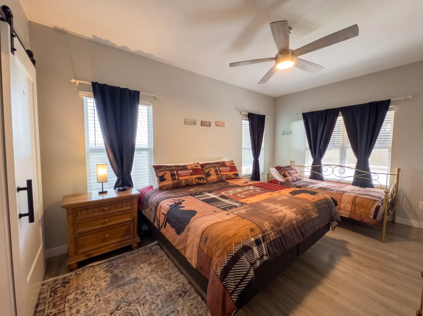 Bedroom with wood-look flooring, a large bed with wildlife-themed bedding, a smaller metal-framed daybed, a wooden nightstand, and windows with dark blue curtains.