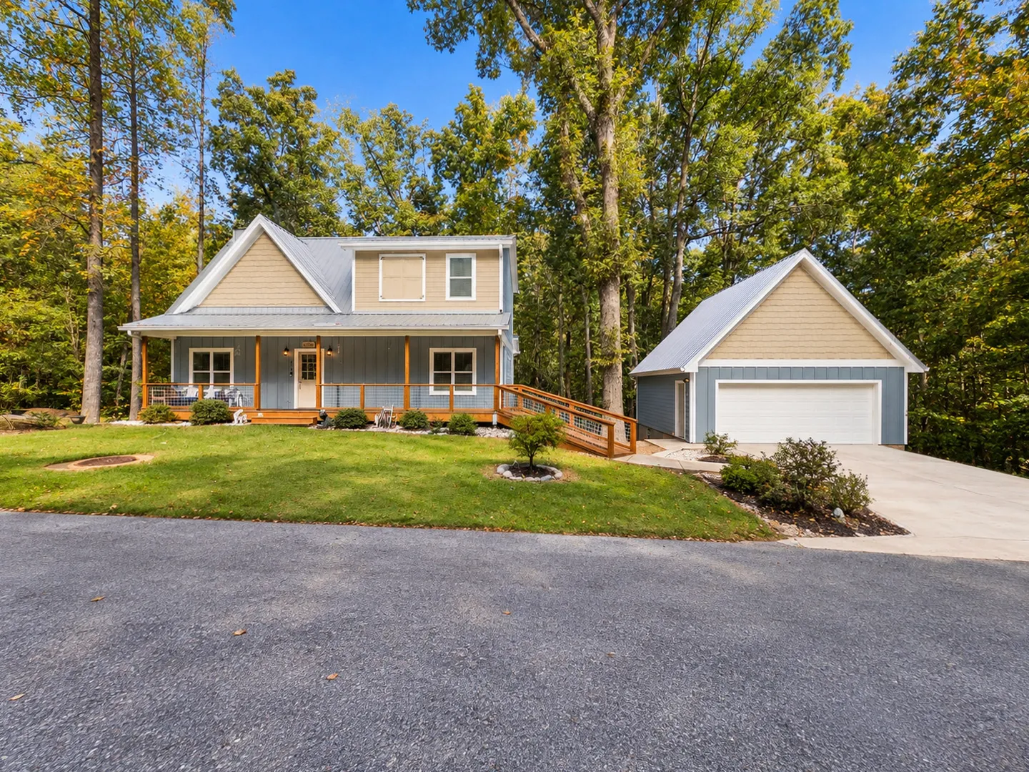 Exterior view of a blue and beige house with a front porch, ramp, and detached garage surrounded by trees.