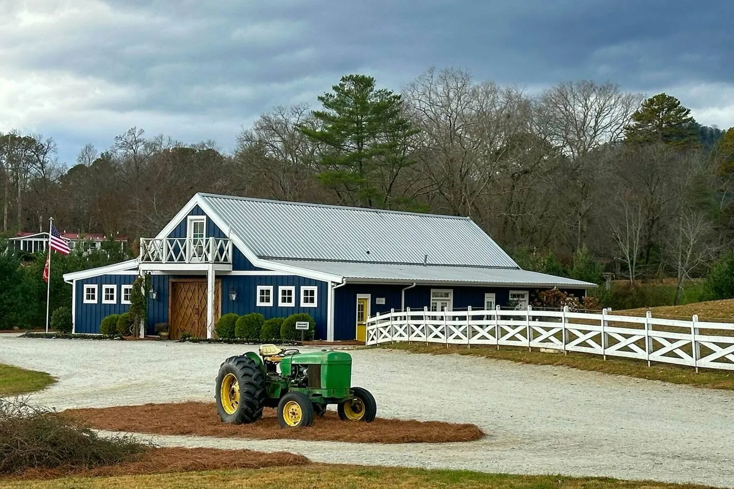 A blue barn-style building with a metal roof and white trim, featuring an upper balcony, white fencing, and a green vintage tractor on the gravel driveway.