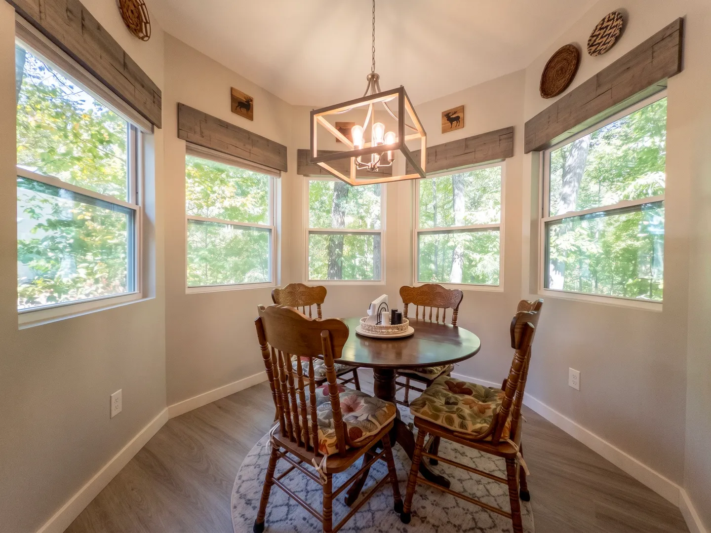 Dining nook with a round wooden table, four wooden chairs, and windows overlooking green trees.