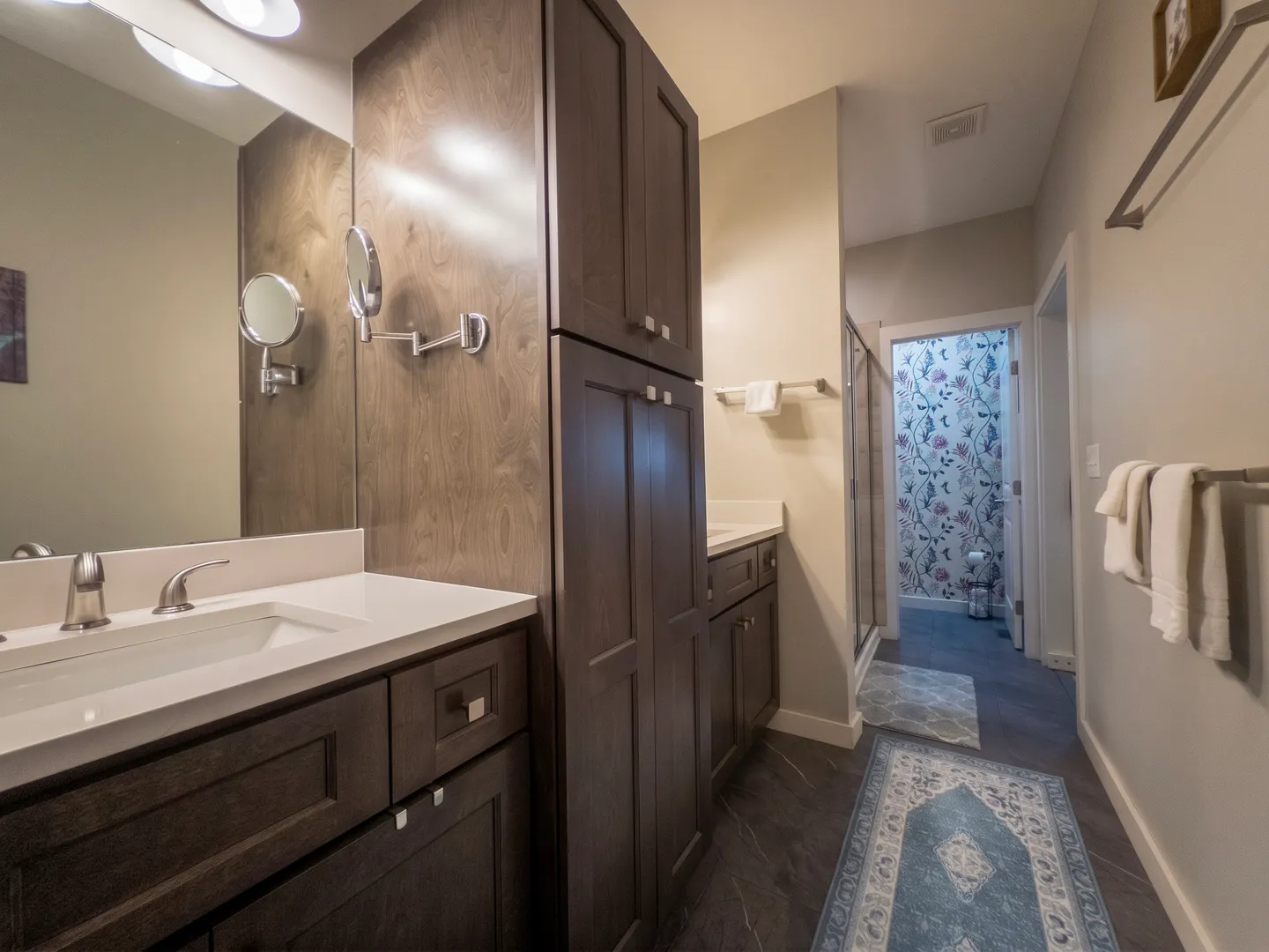 Bathroom vanity area with dark wood cabinets, white countertops, tall storage cabinet, and a view into a patterned wallpaper room.