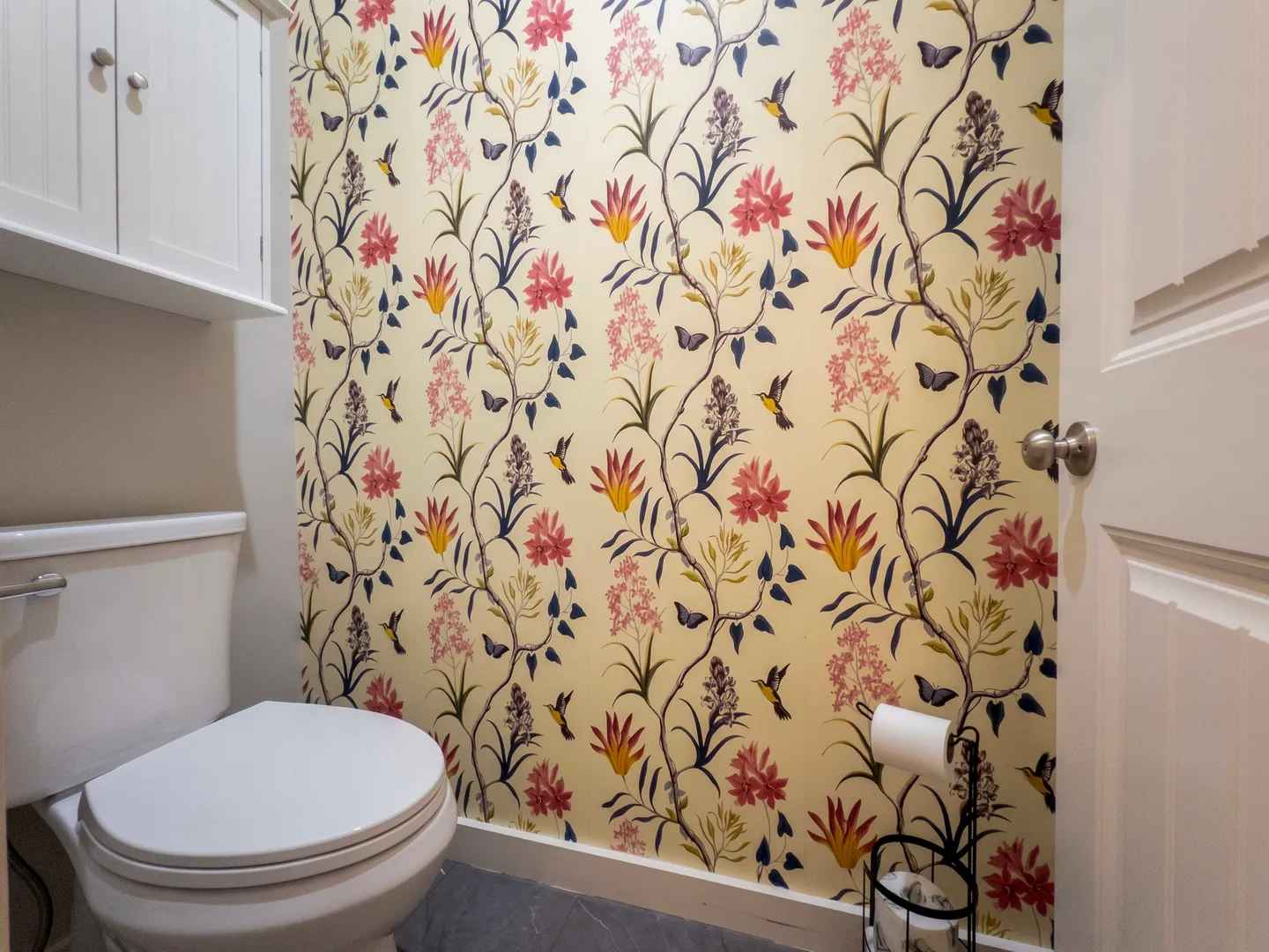 Bathroom with a toilet, floral and bird pattern wallpaper, and a white upper cabinet.