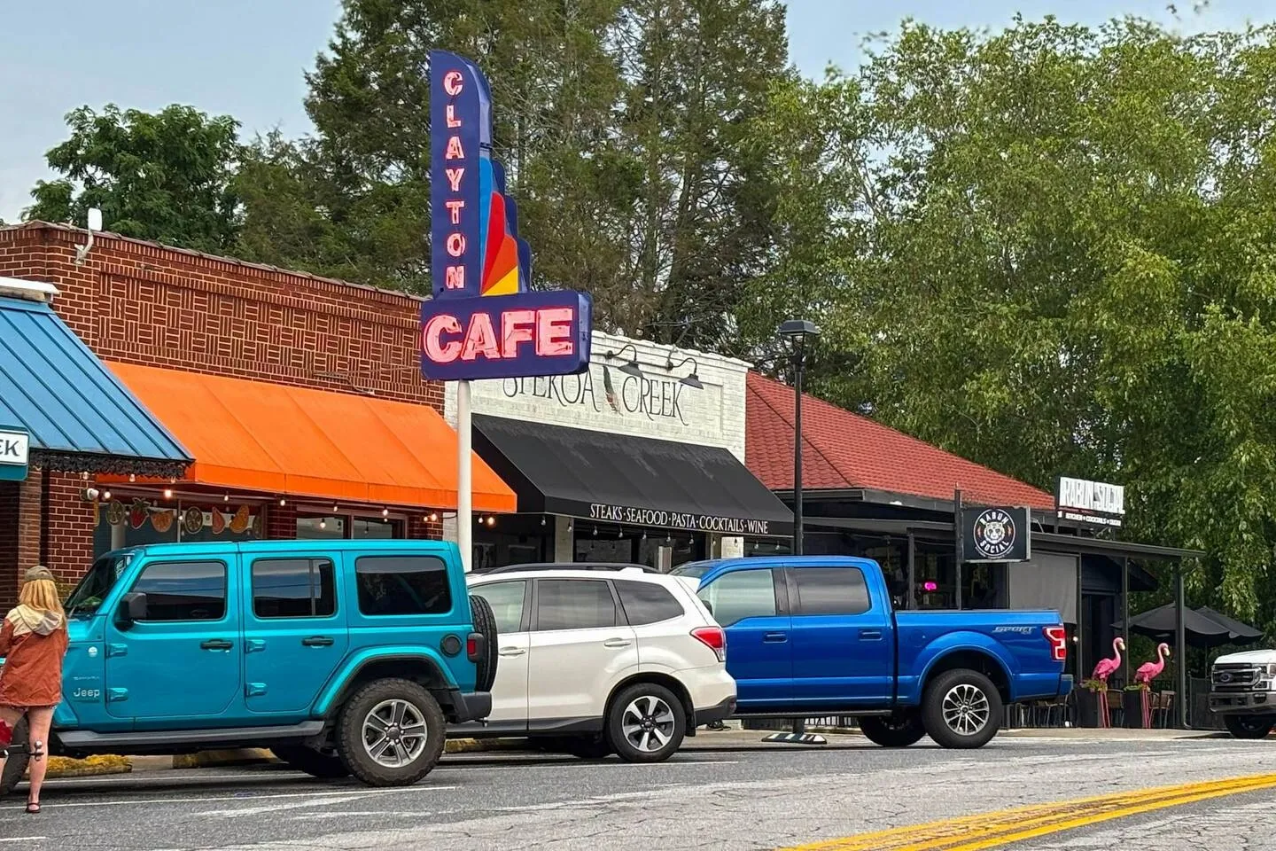 Street view of local storefronts including Clayton Cafe with parked cars along the curb.