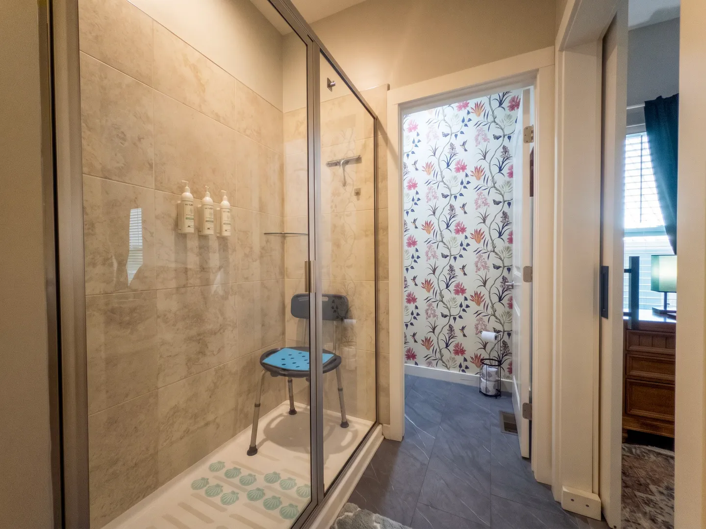 Bathroom featuring a glass-enclosed walk-in shower with a shower chair, leading into a water closet with floral wallpaper.