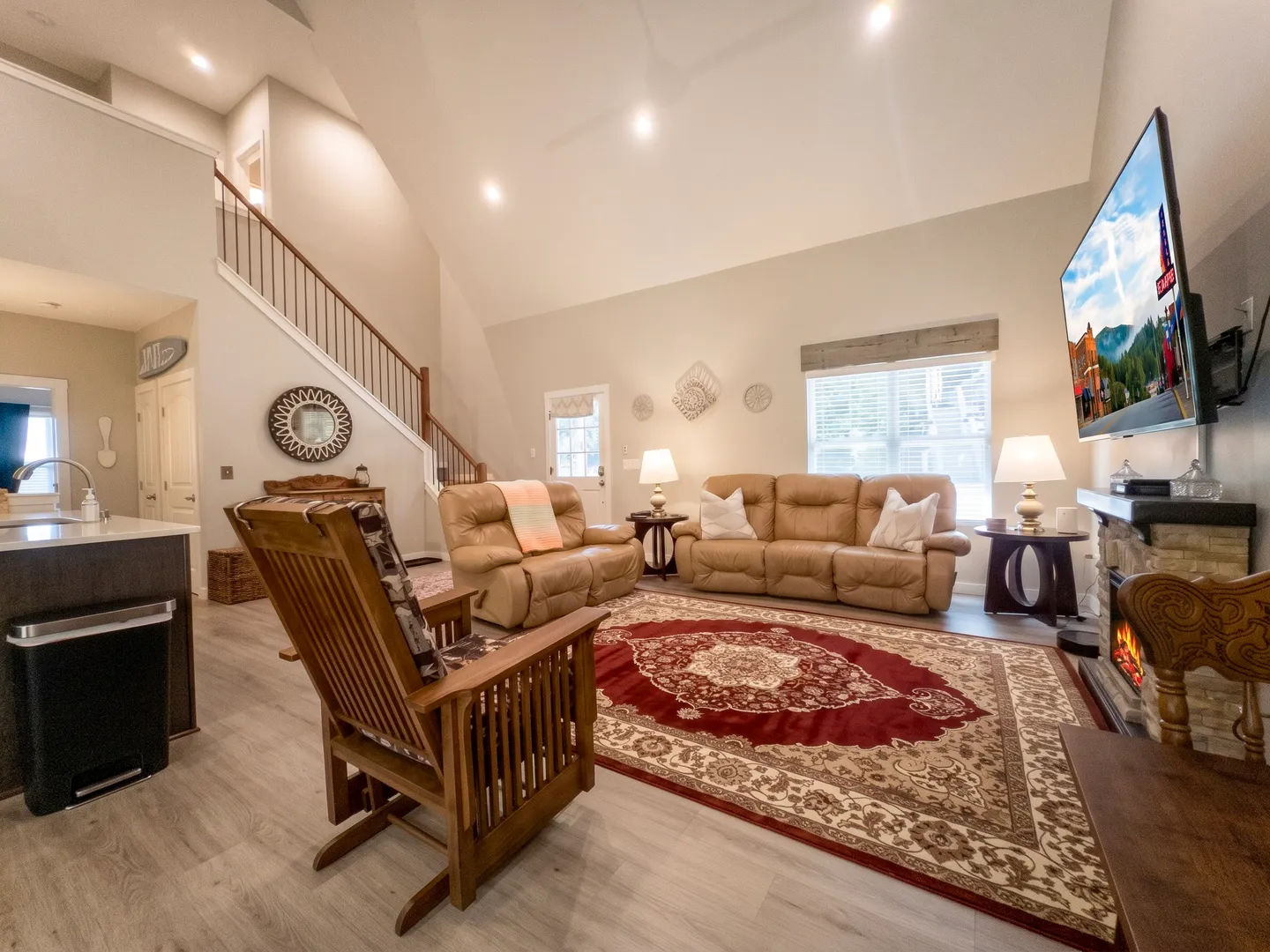 Open-concept living room with vaulted ceiling, leather seating, a rocking chair, area rug, fireplace, TV, and staircase in the background.