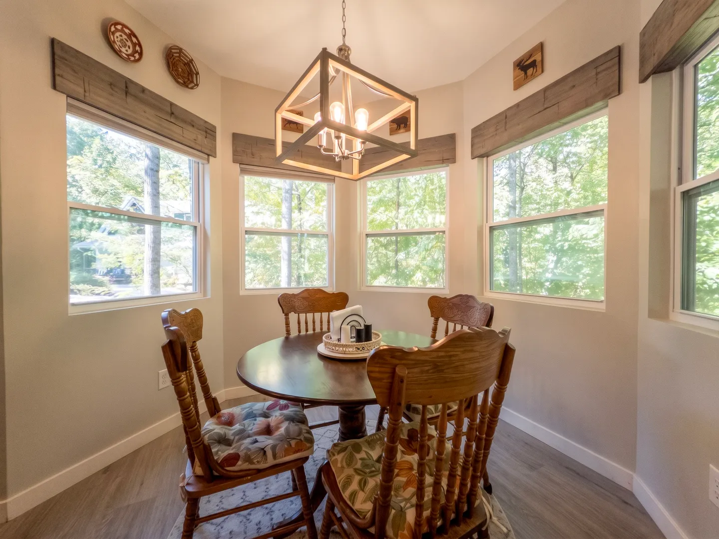 Dining nook with a round wood pedestal table, four wooden chairs, and large windows looking out onto green trees.