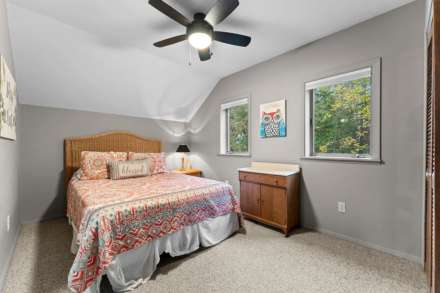 Bedroom with a patterned bedspread, wicker headboard, sloped ceiling, ceiling fan, and two windows.