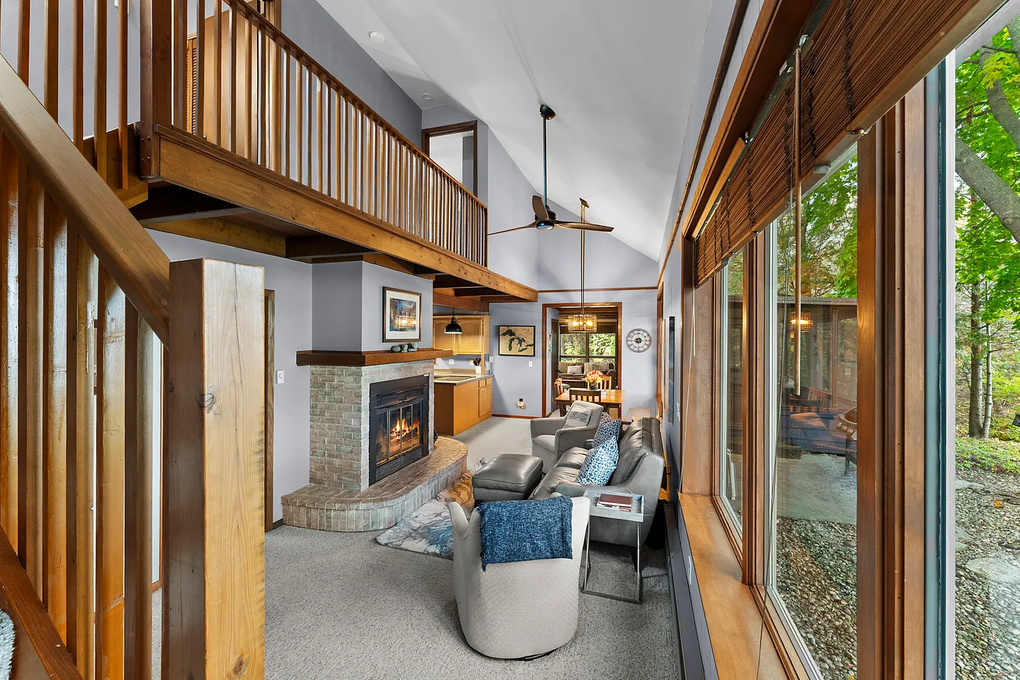 Living room with vaulted ceiling, wood-trimmed fireplace, carpeted flooring, large picture windows, and an overlooking loft.