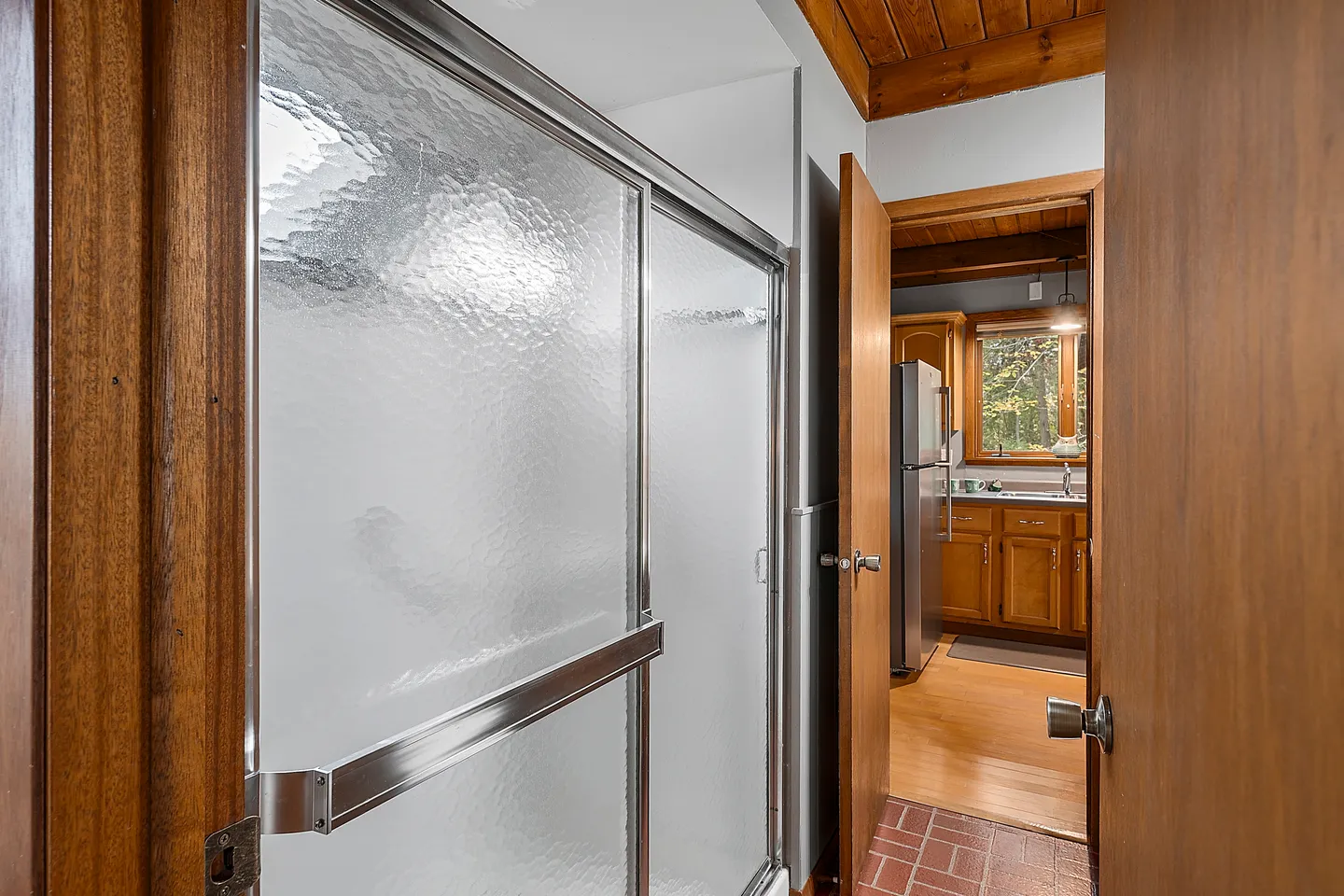 Bathroom with a frosted glass shower door opening into a hallway that leads to a kitchen.