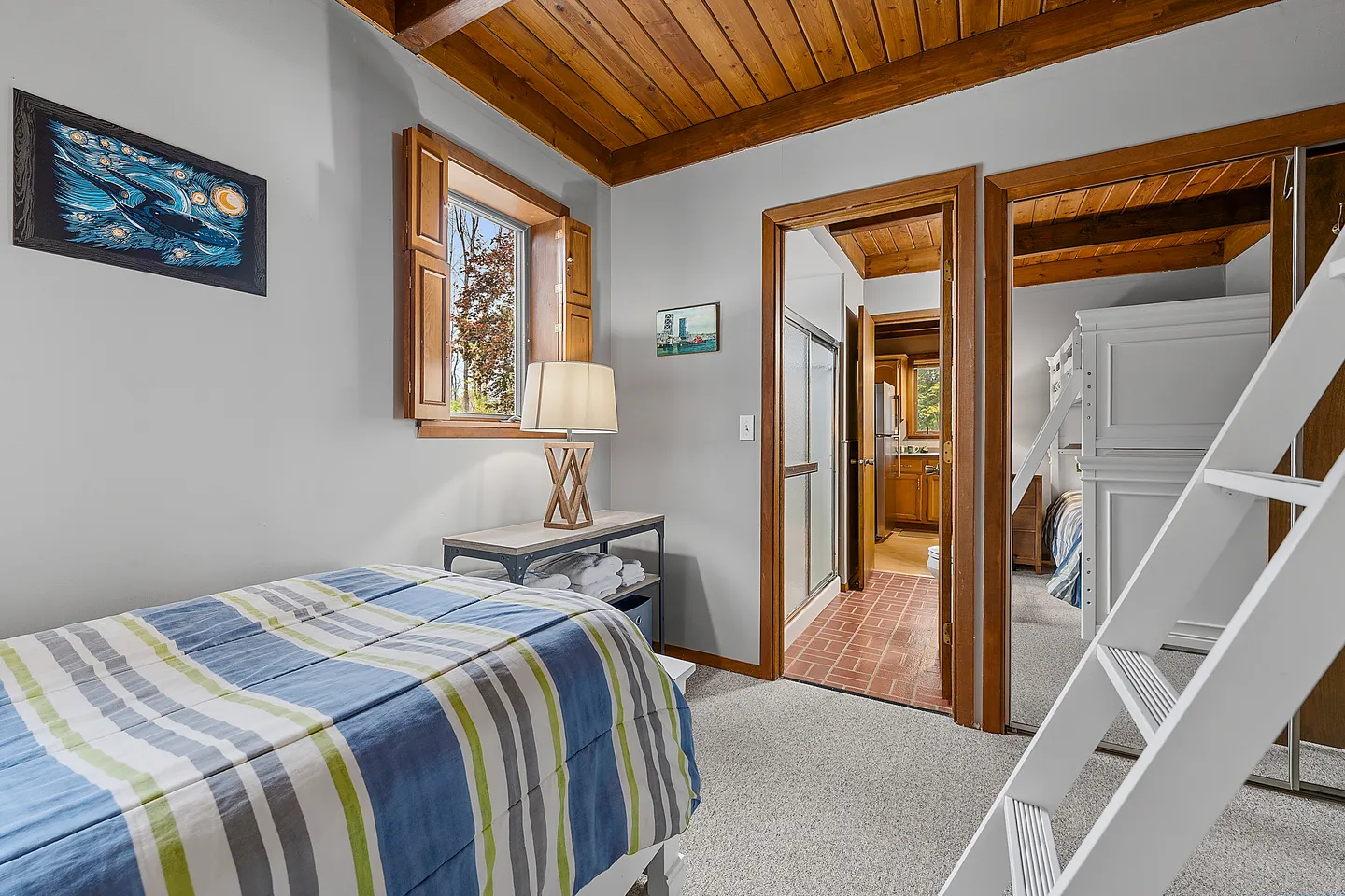 Bedroom with a single bed featuring striped bedding, wood-plank ceiling, and doorways showing an adjacent bathroom and another room reflected in mirrored closet doors.