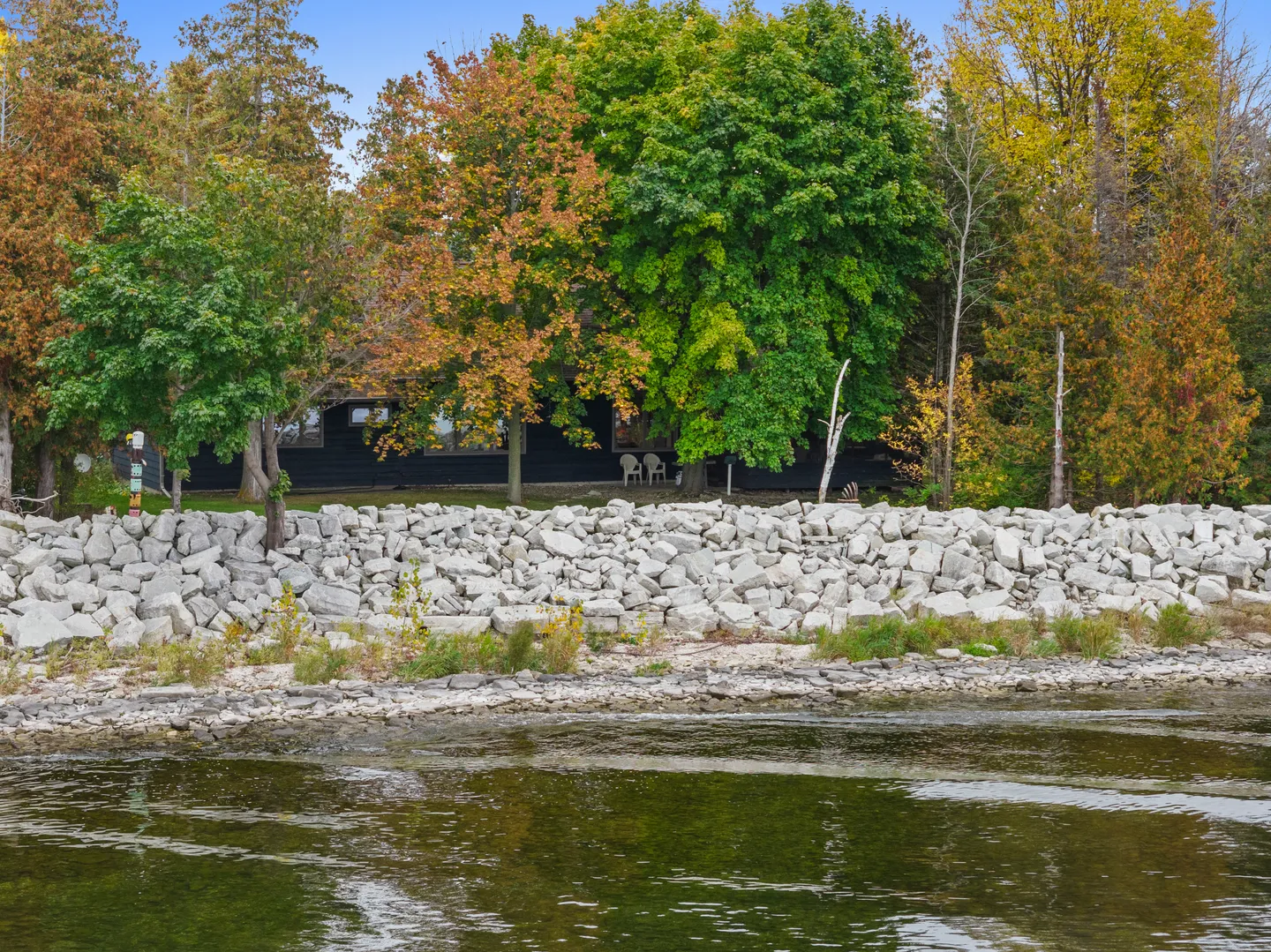 View from the water of a dark-sided cabin nestled behind a rock seawall and surrounded by fall foliage.