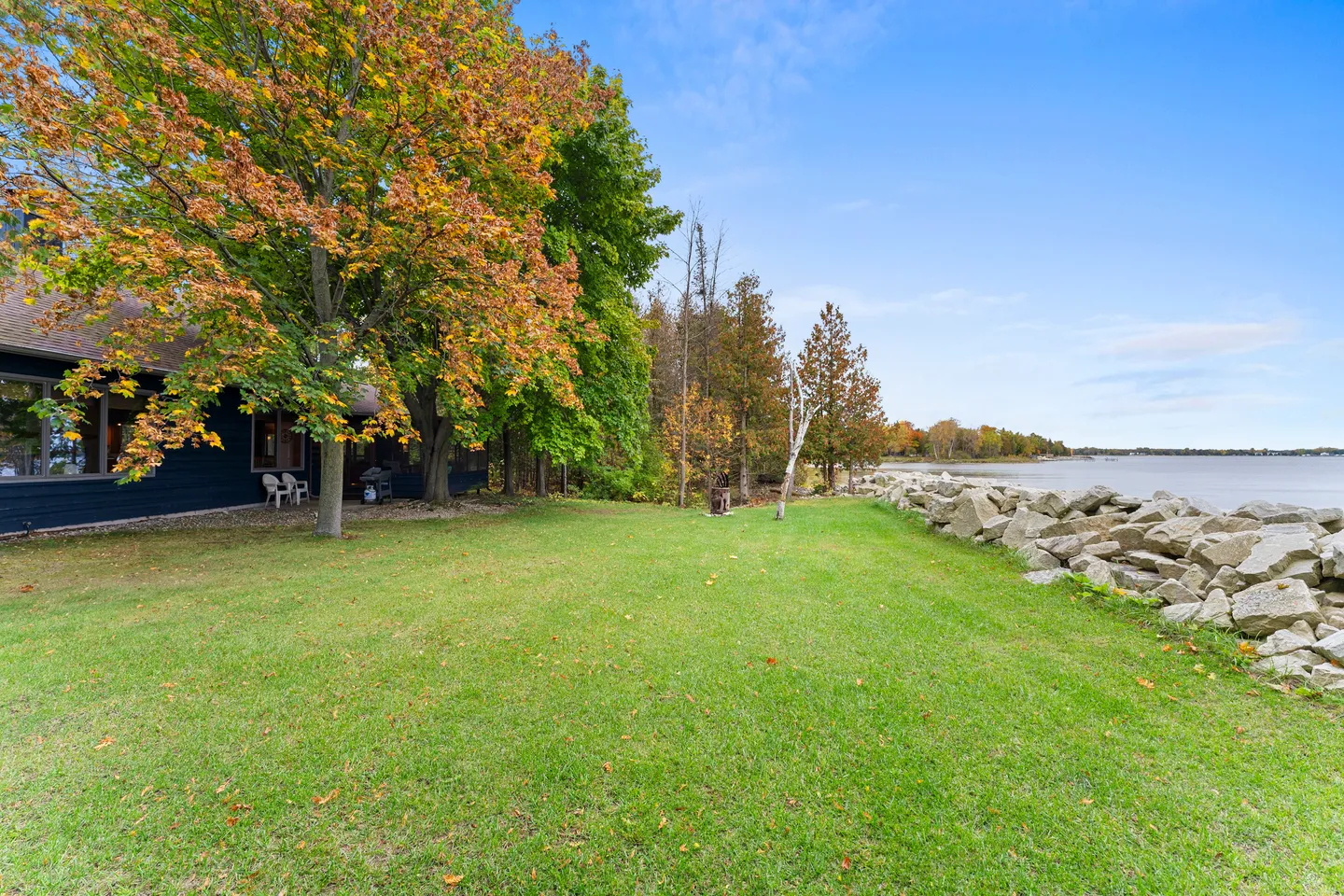 Grassy waterfront yard bordered by trees and large shoreline rocks under a bright blue sky.