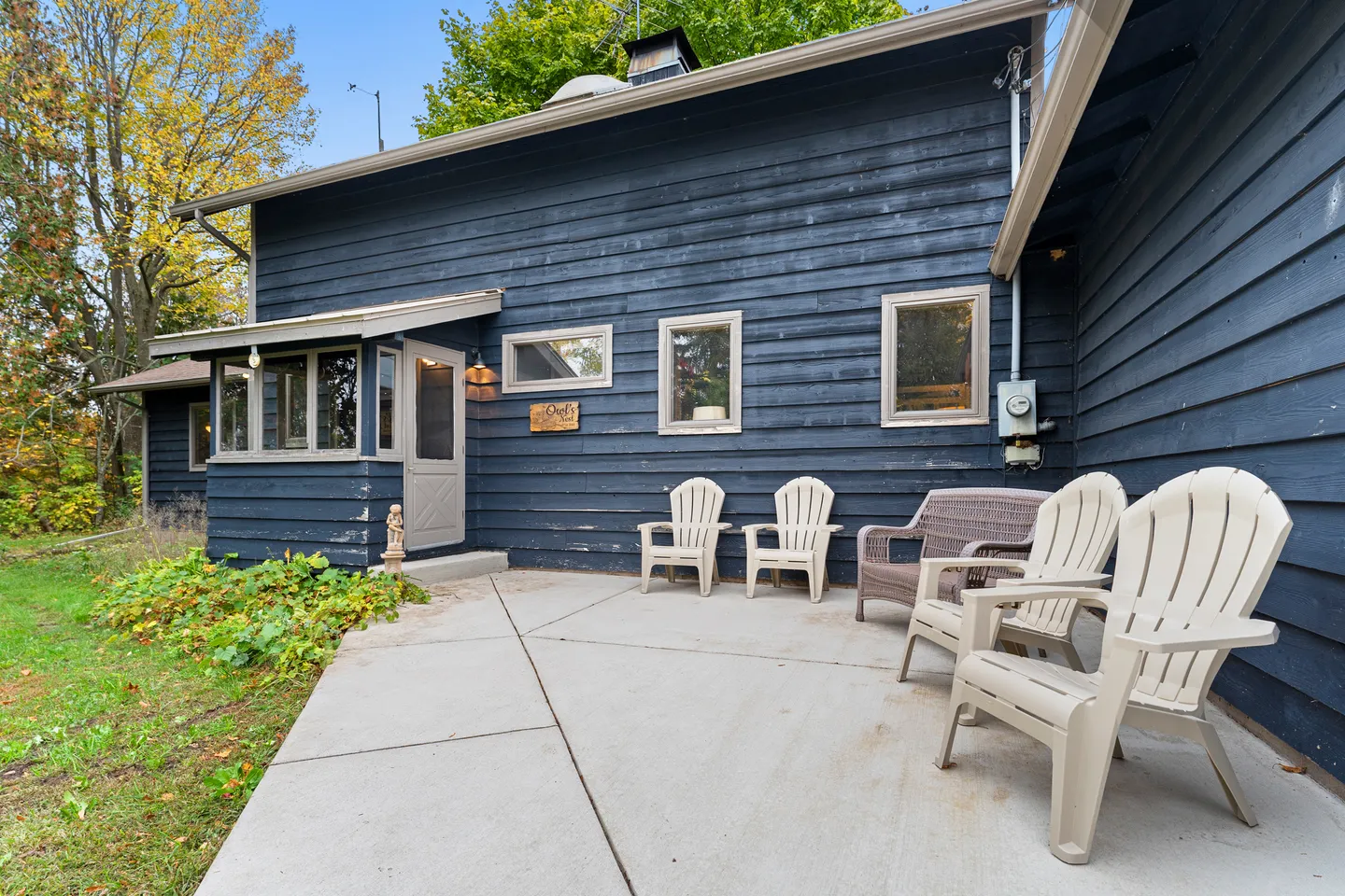 Concrete patio with Adirondack chairs outside a dark blue horizontal-sided house with autumn trees in the background.