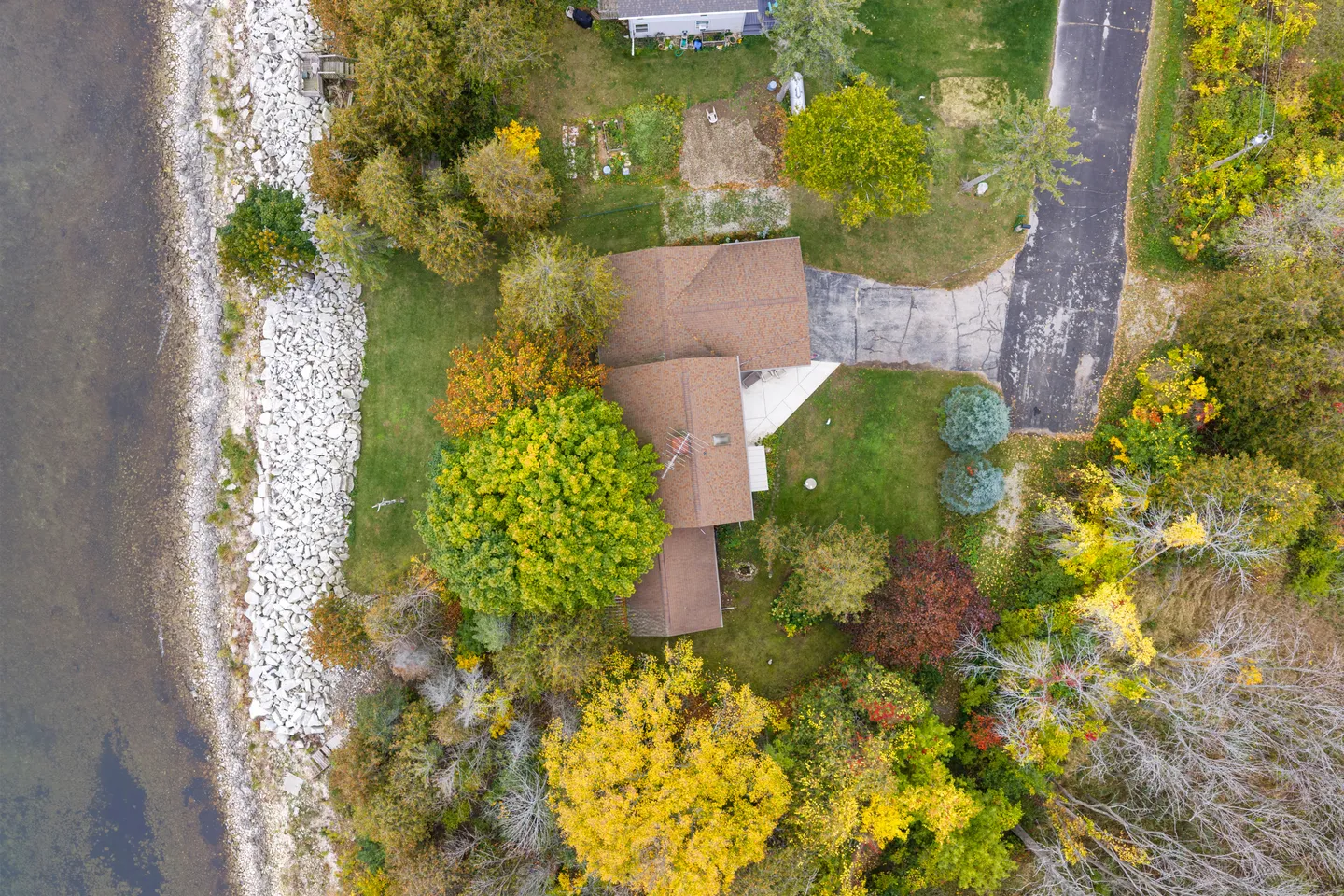 Overhead aerial view of a lakeside home surrounded by fall-colored trees, a rocky shoreline, and an asphalt driveway.