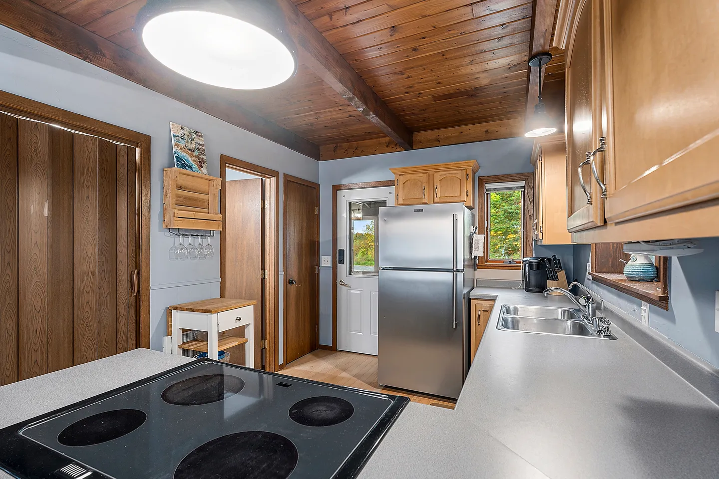 Kitchen interior showing a cooktop in the foreground, stainless steel refrigerator, sink, and wood plank ceiling.