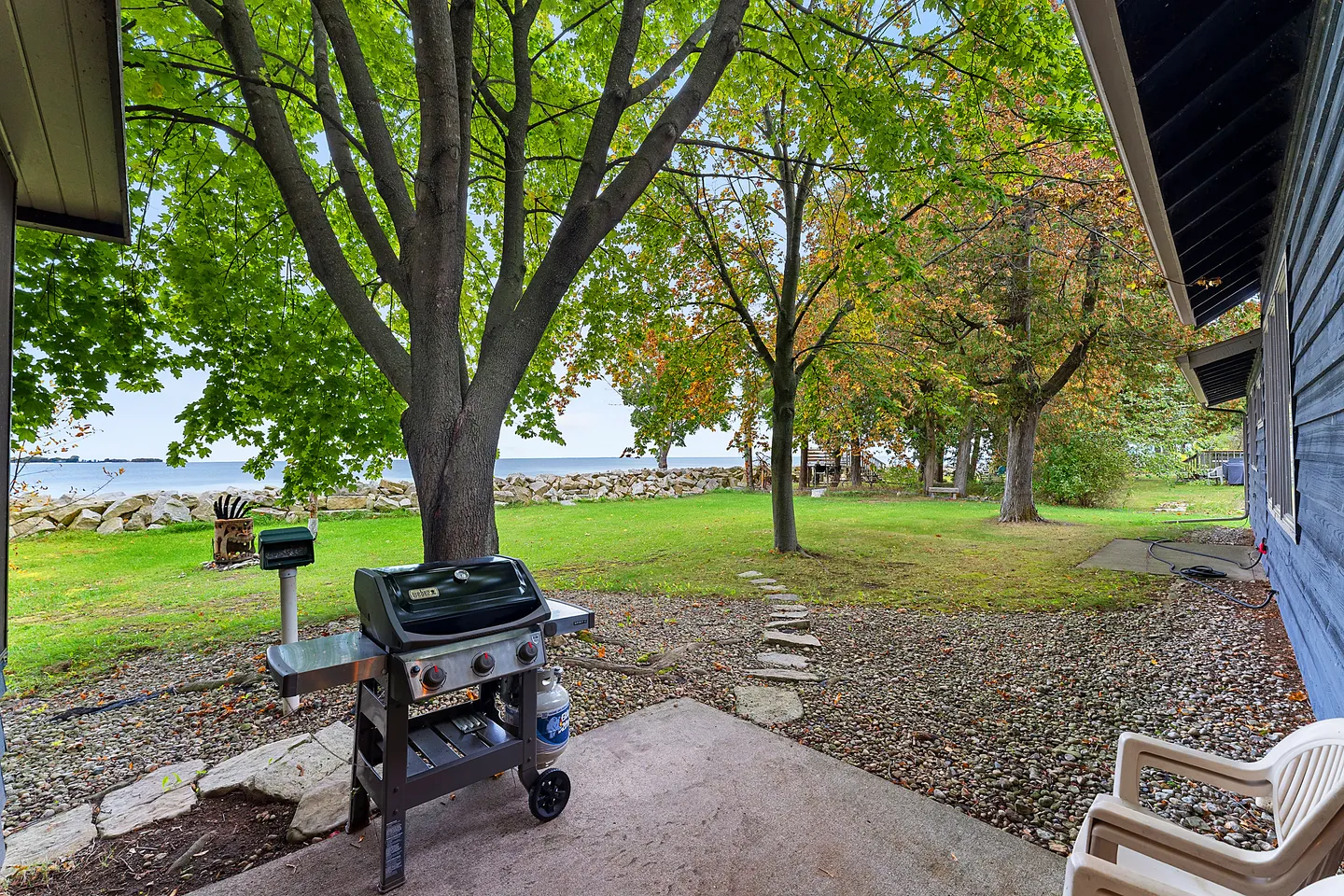 A backyard view from a concrete patio showing a barbecue grill, large shade trees, a stone pathway, and a body of water in the distance.