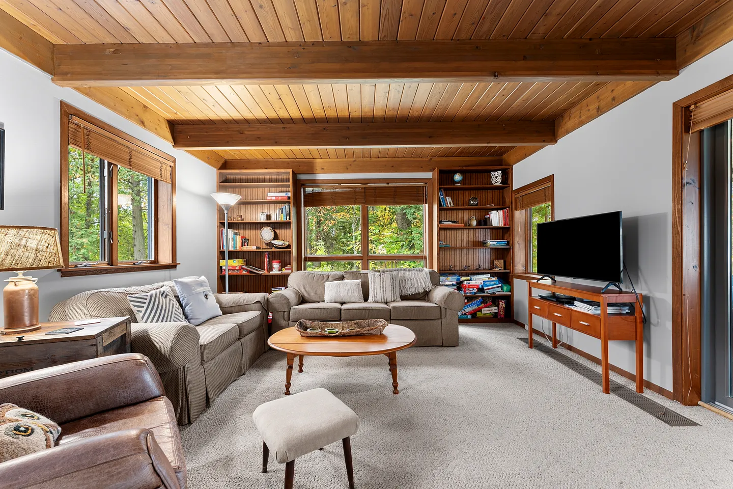 Living room with wood-plank ceiling, exposed beams, carpeted floor, couches, bookshelves, and a TV on a stand.