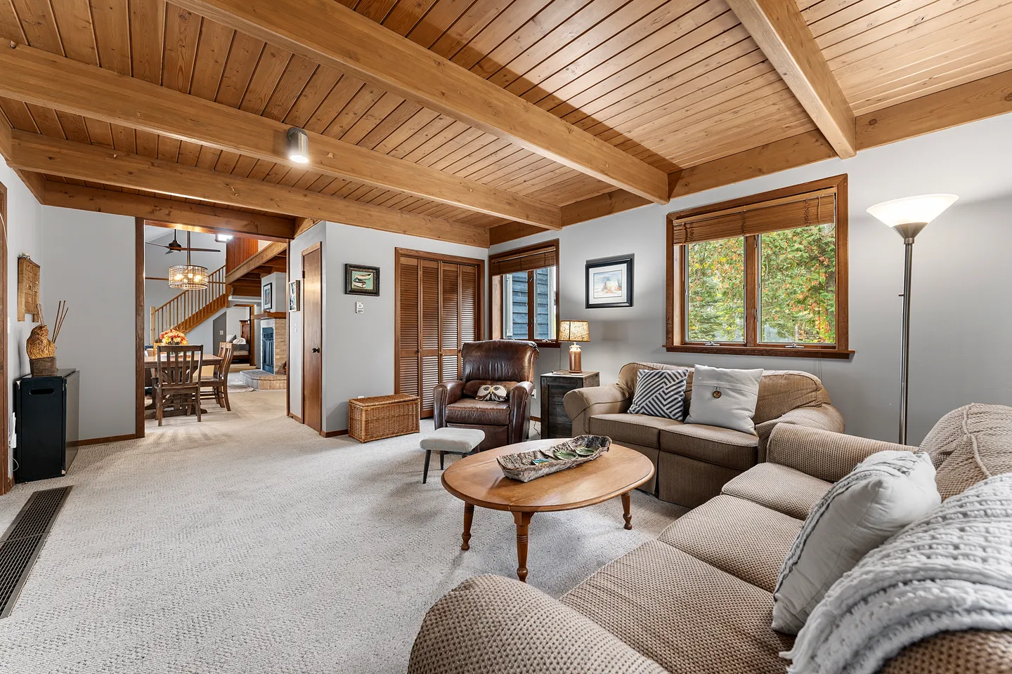 Living room with wood-paneled ceiling, exposed beams, carpeting, neutral upholstered sofas, and an oval coffee table.