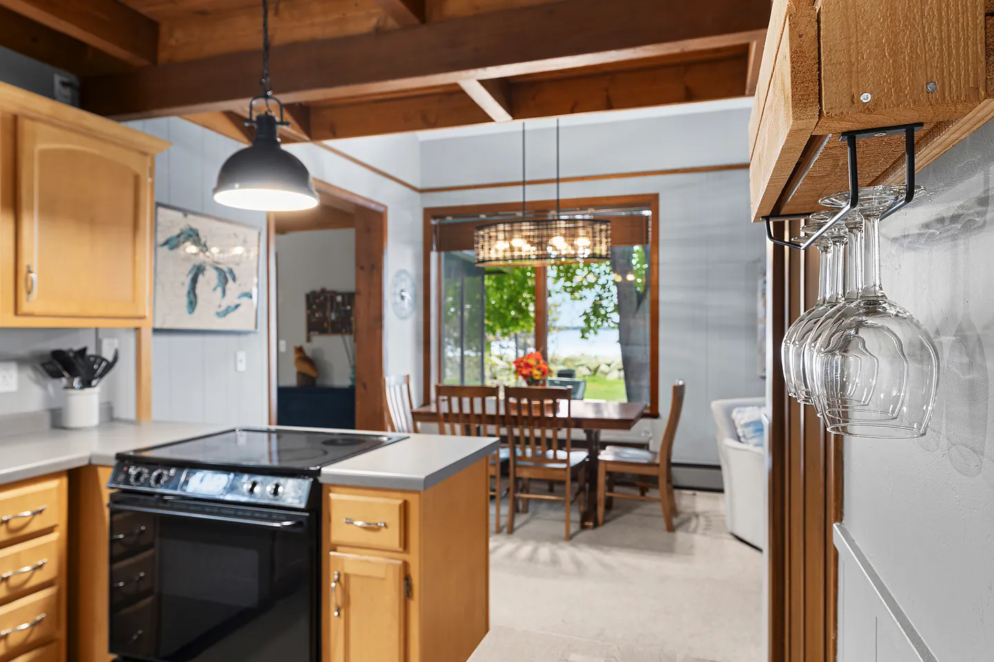 View from a kitchen counter with an electric range toward an adjacent dining area with a wooden table, chairs, and large window overlooking the water.