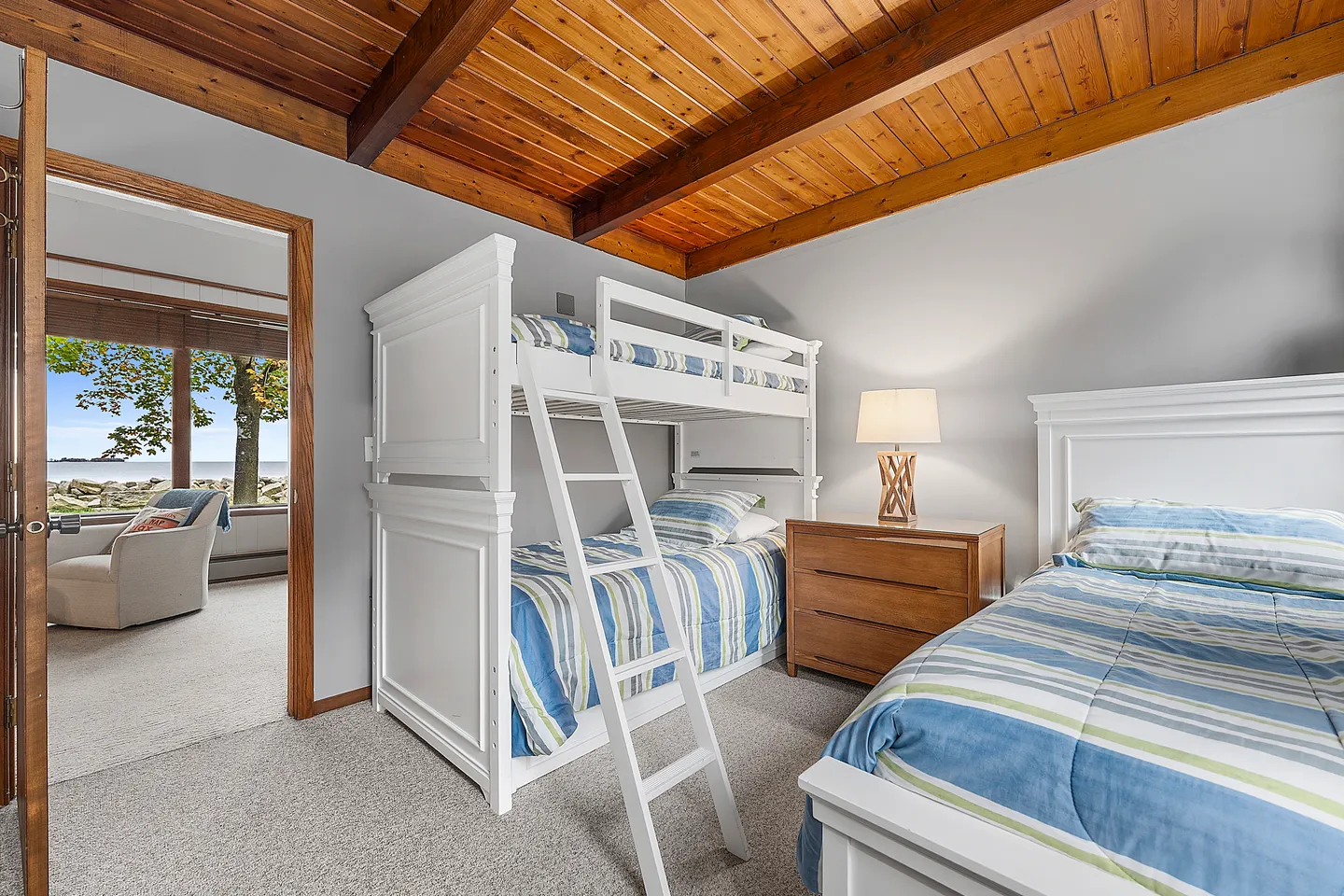 Bedroom featuring a white bunk bed, a single bed, a wooden nightstand, and an angled wood-plank ceiling with exposed beams.