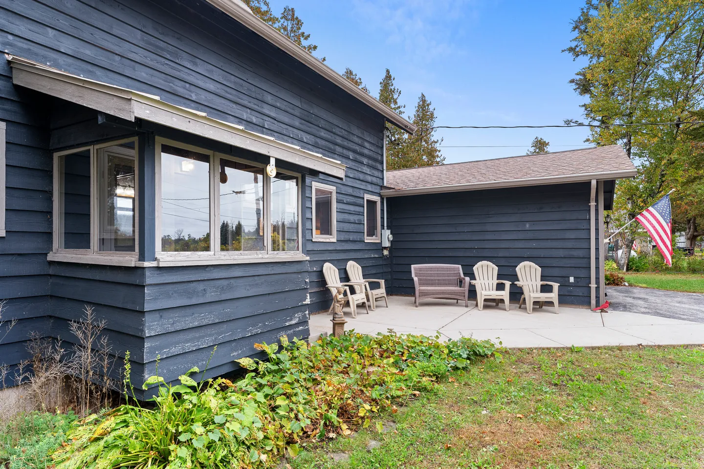 Exterior view of a dark blue wood-sided house with a concrete patio featuring outdoor chairs and a bench next to a lawn and garden bed.