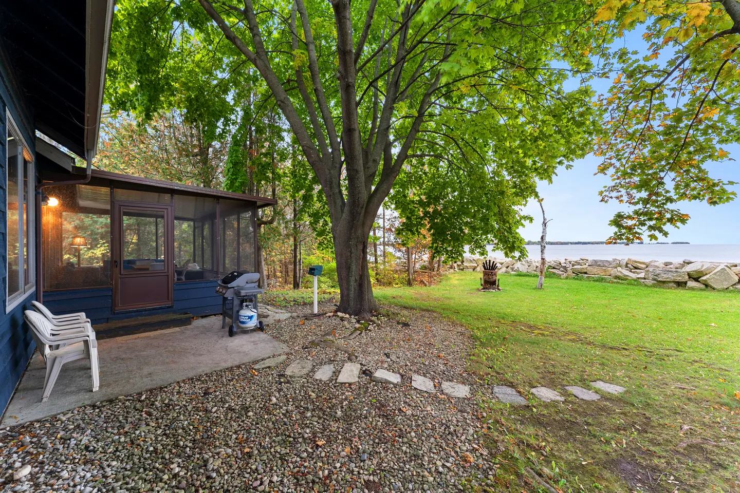 Exterior view of a blue home with a screened porch, gas grill, lawn, mature trees, and a lakefront shoreline.