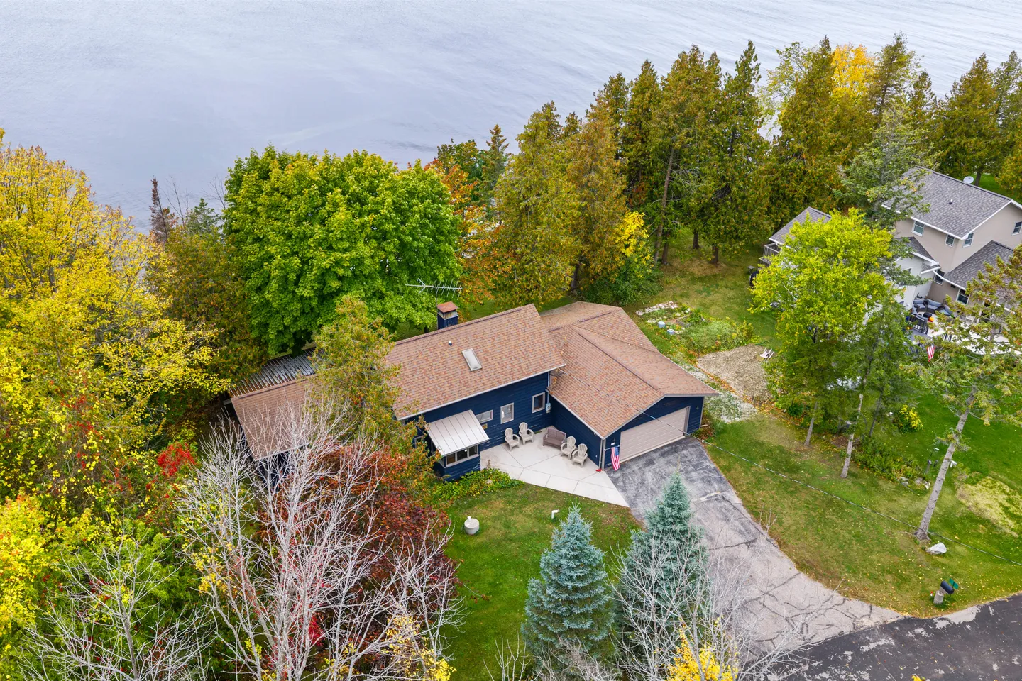 Aerial view of a blue one-story house with a brown roof surrounded by autumn trees on a lake shoreline.