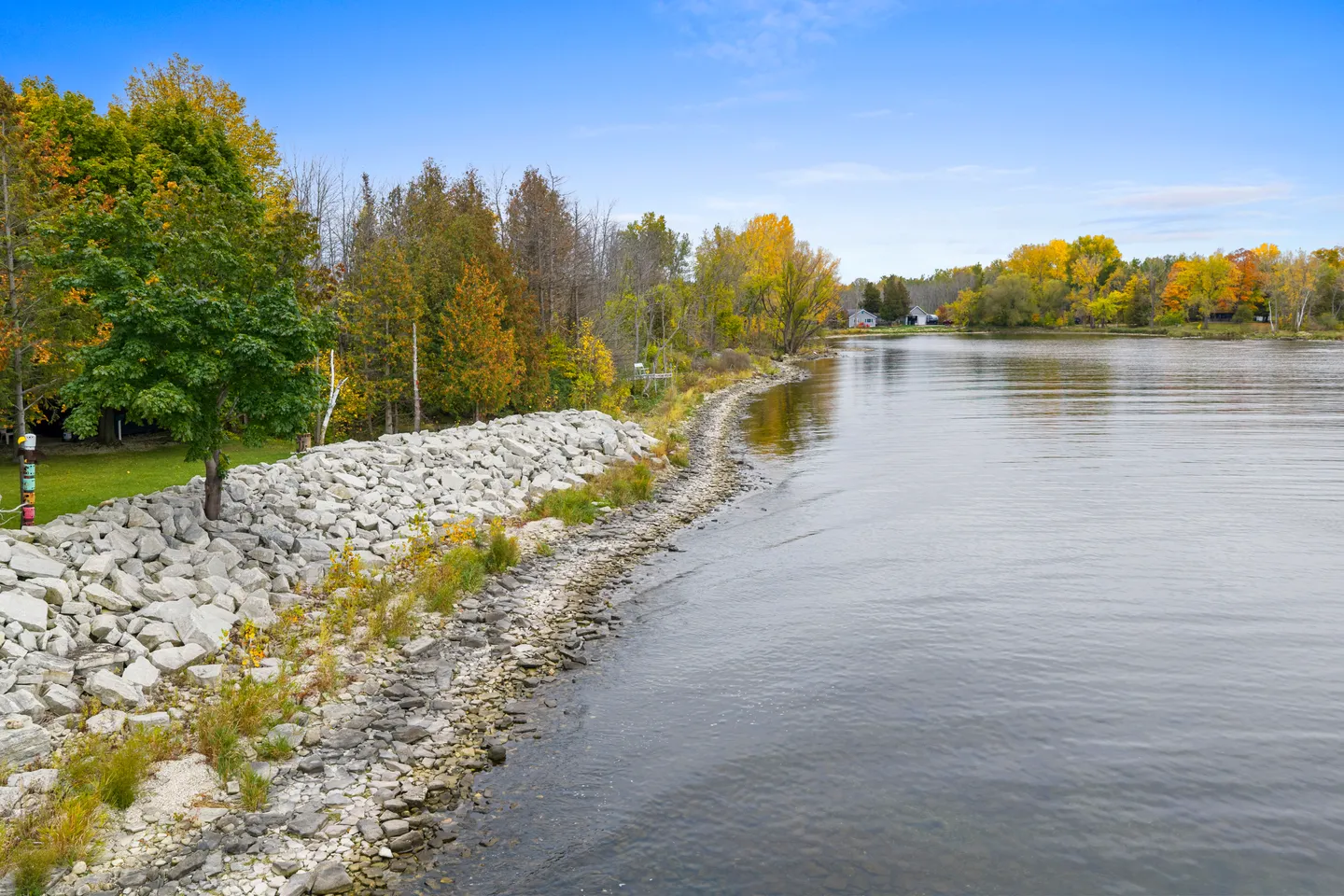 Elevated view of a rocky shoreline along a wide body of water bordered by autumn trees and a blue sky.