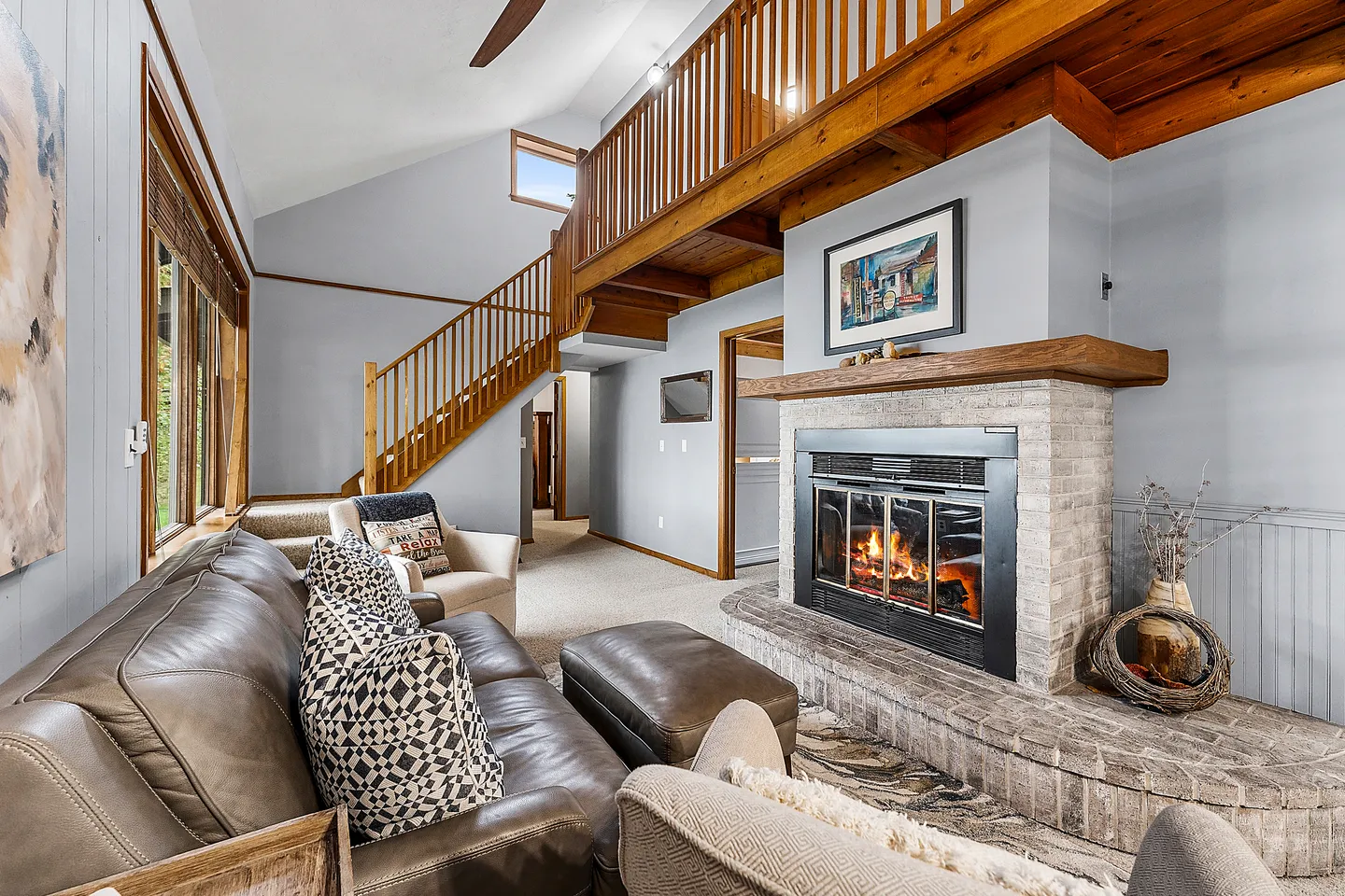 Living room featuring a brown leather sofa, an active brick fireplace with a wood mantel, and an open wooden staircase leading to a loft.
