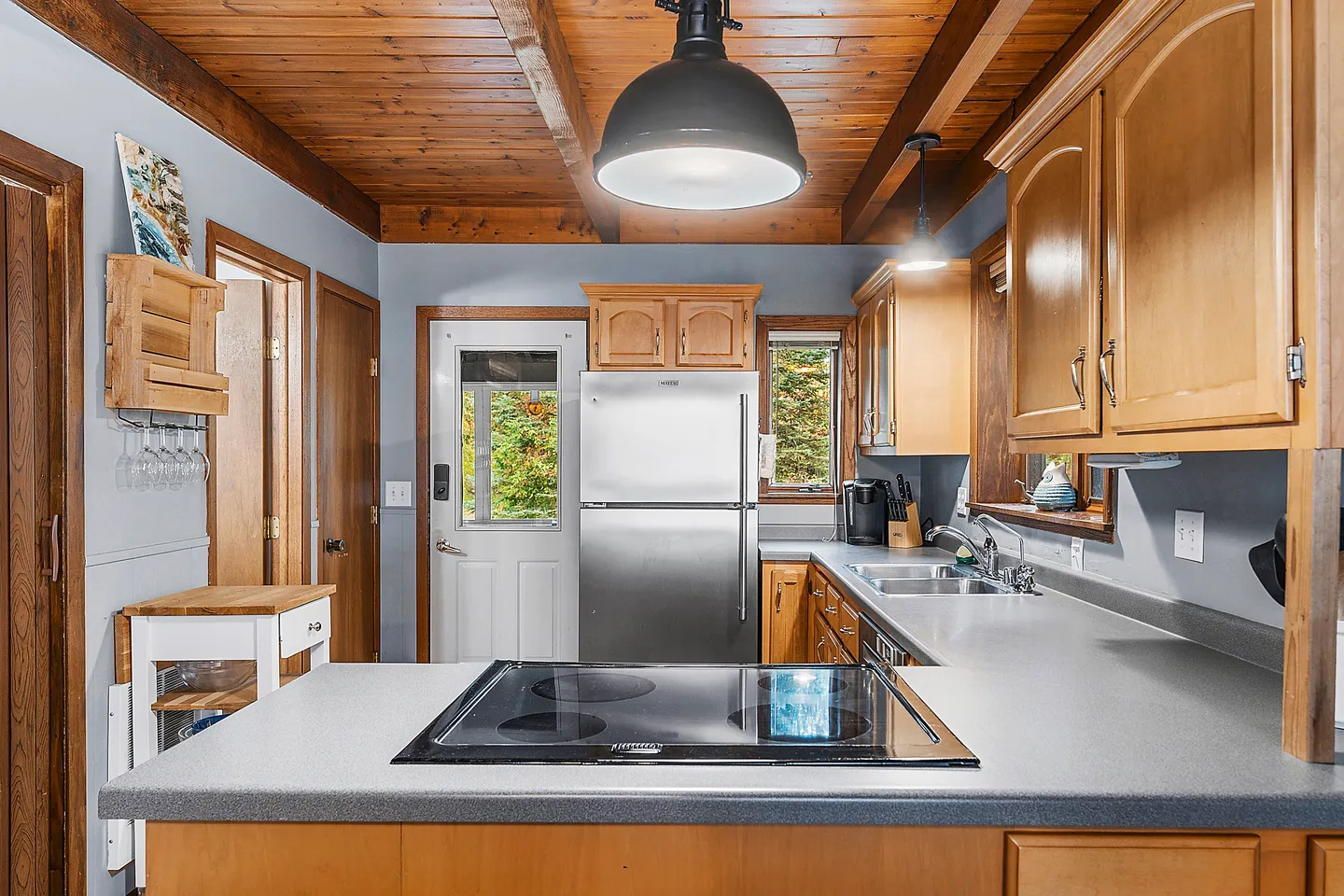 Kitchen interior featuring light wood cabinets, gray countertops with an electric cooktop, stainless steel refrigerator, and wood plank ceiling.