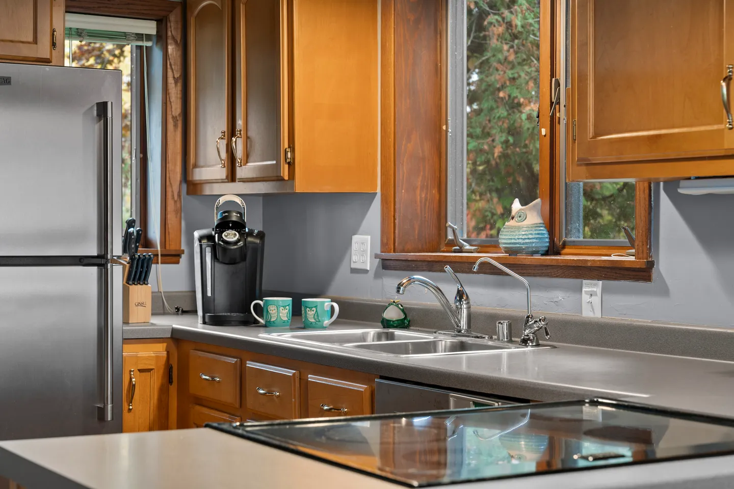 Close-up view of a kitchen counter with a double stainless steel sink, wood cabinetry, refrigerator, coffee maker, and a window.