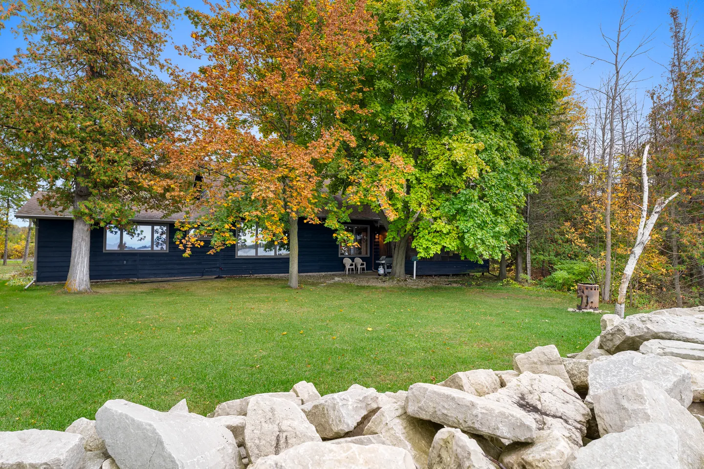 Exterior view of a dark-sided home behind a grassy lawn, autumn trees, and a foreground rock wall.